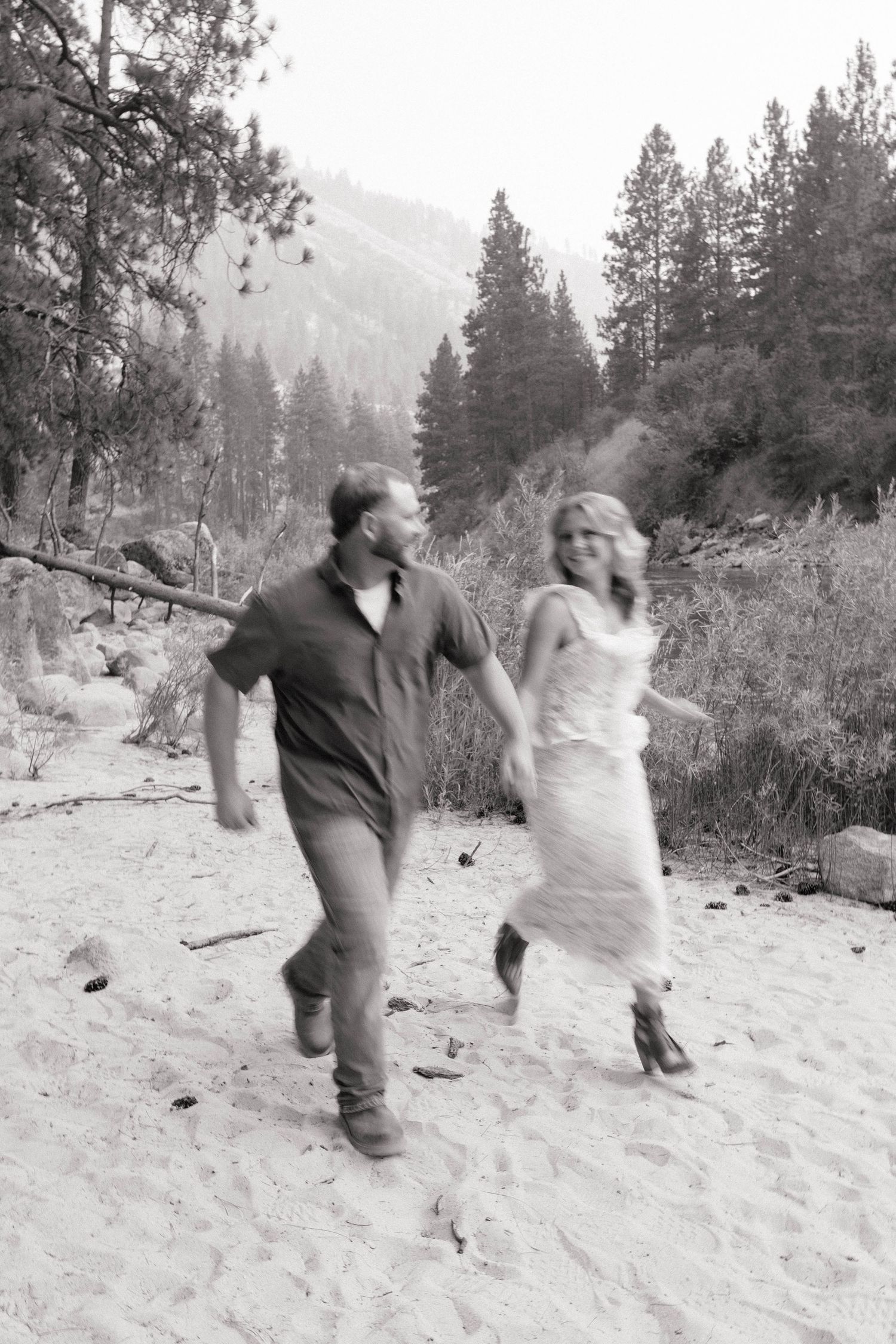 Black and white photo of a couple running hand in hand along a sandy riverbank with evergreen trees and hazy mountains behind them.