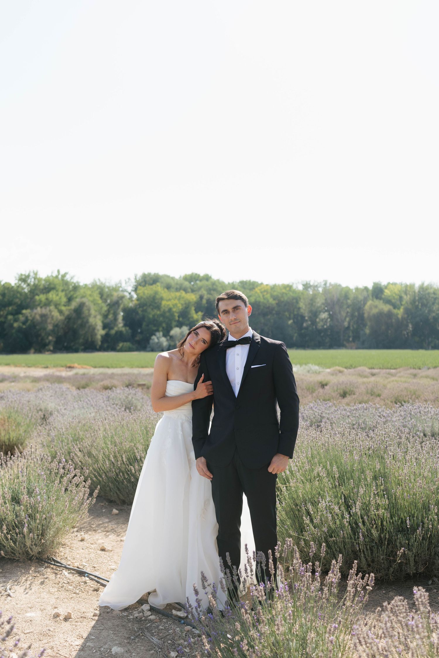 A bride in a white gown leans on a groom in a black tuxedo, posing in a lavender field.