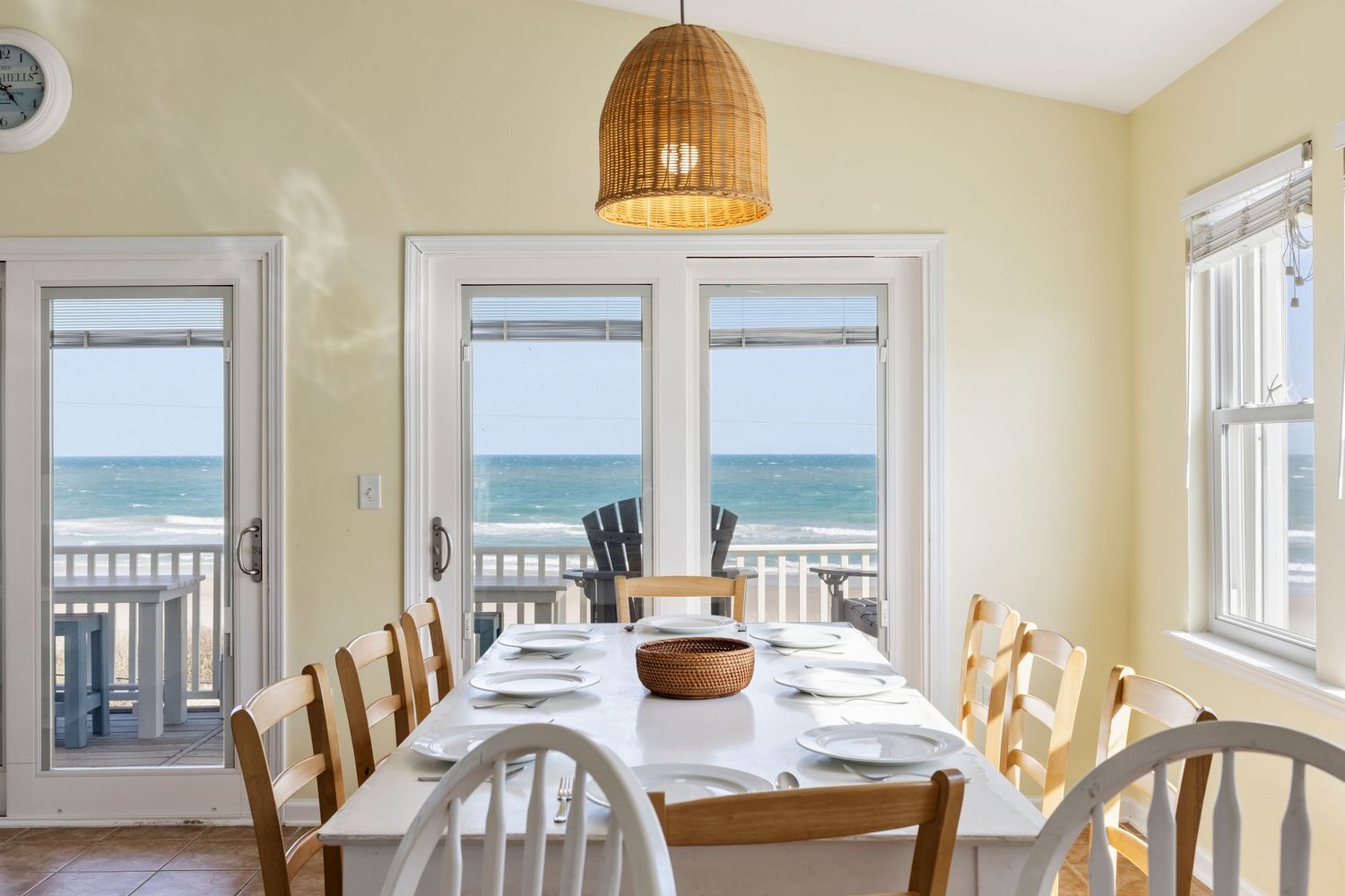 Bright beachfront dining room with white table, wicker chairs, and panoramic ocean views through French doors.