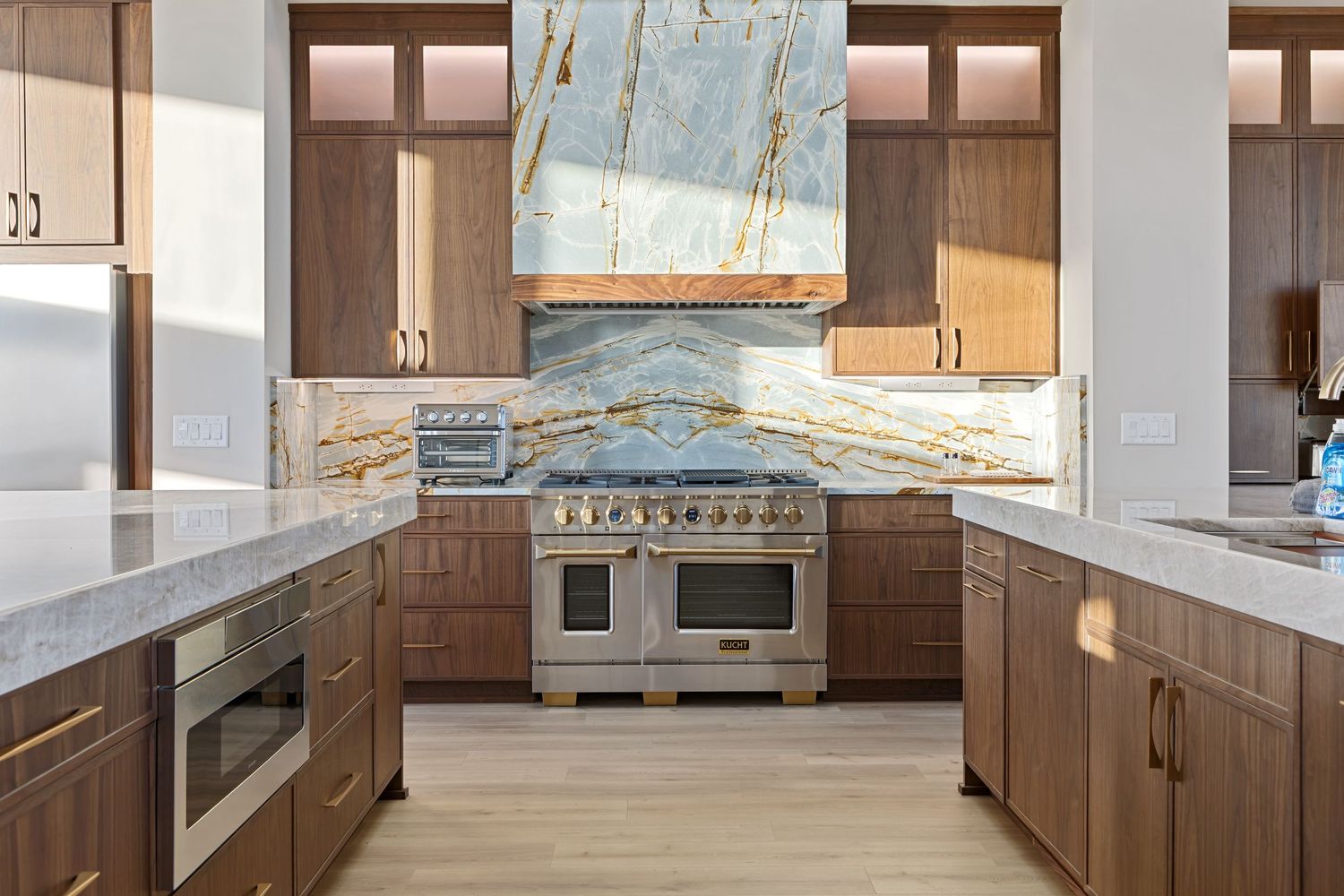 Modern kitchen with walnut cabinets, marble backsplash, stainless steel range, and white quartz countertops on light wood floors.