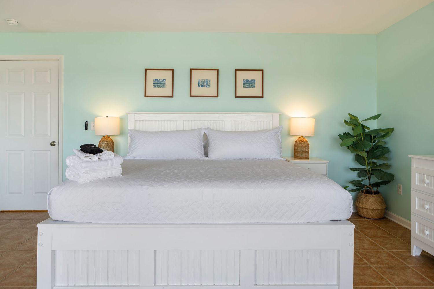 Bright beach-themed bedroom with king bed, white bedding, mint green walls, framed art, and a tropical plant.
