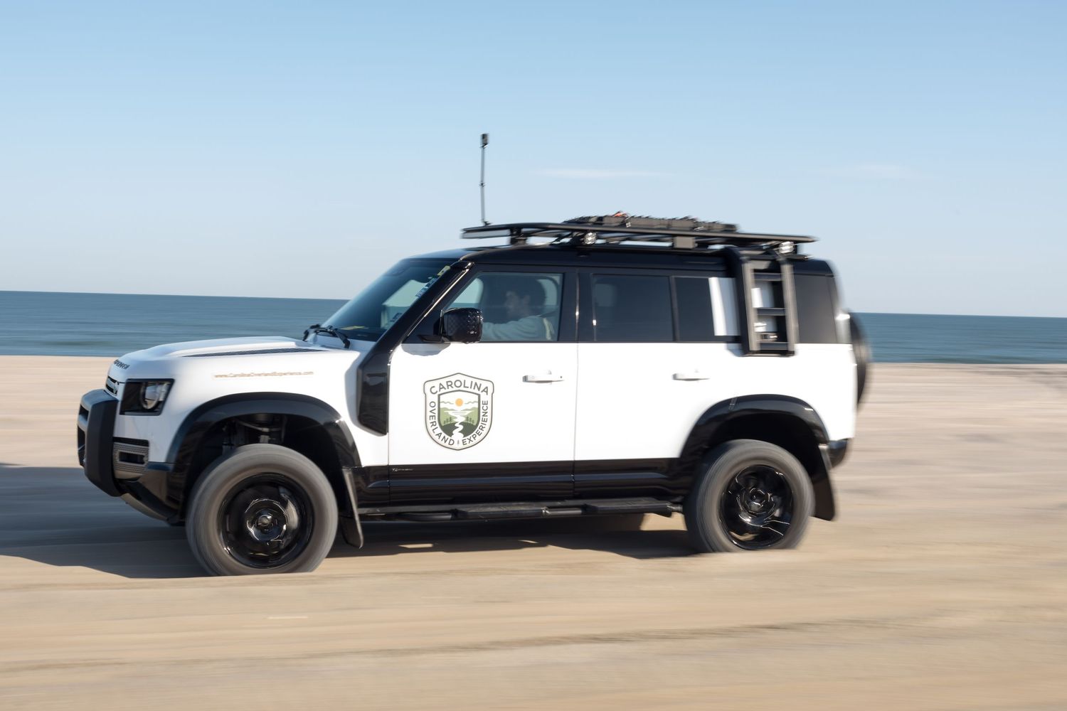White off-road vehicles driving on a sandy beach with ocean in the background.