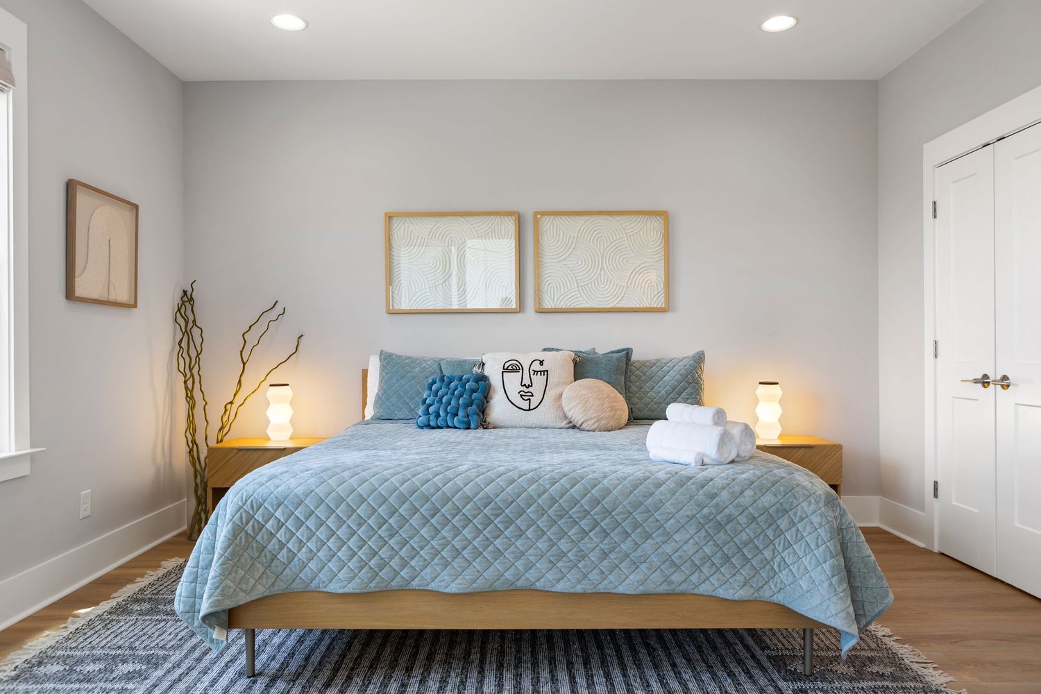 Modern bedroom with blue quilted bedspread, wooden nightstands, framed wall art, and soft neutral tones.