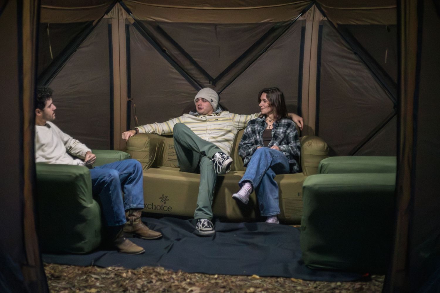 Three people relaxing on green sofas inside a canvas tent shelter during an outdoor camping trip.