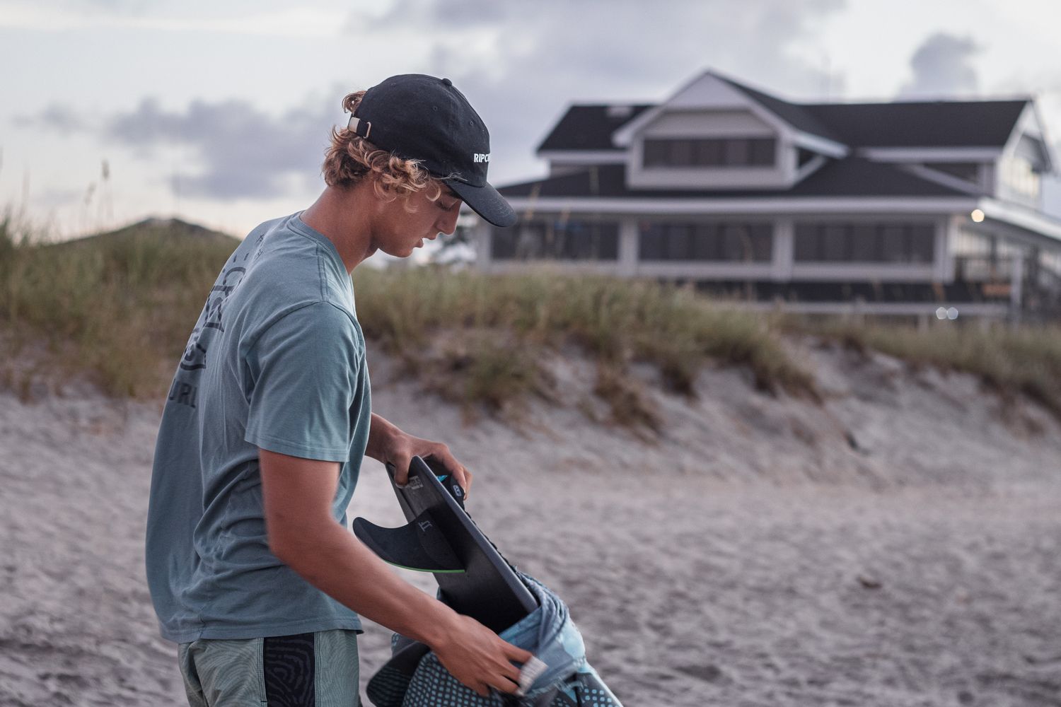 Surfer preparing equipment on a sandy beach with beachfront houses in the background.