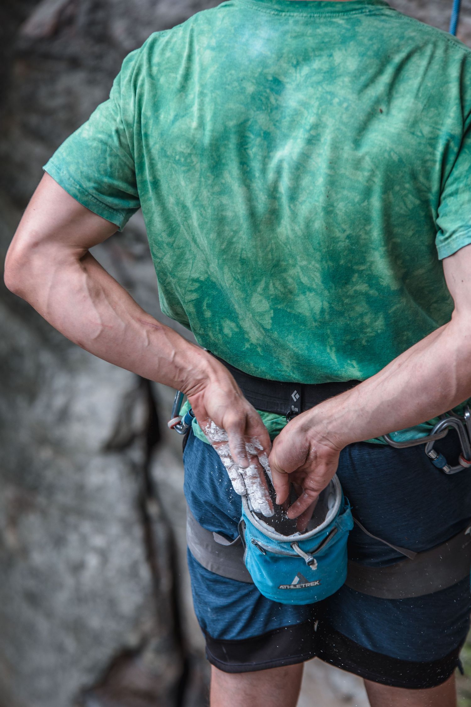 Close-up of a rock climber's chalk bag and harness against a green shirt, with rock face in the background