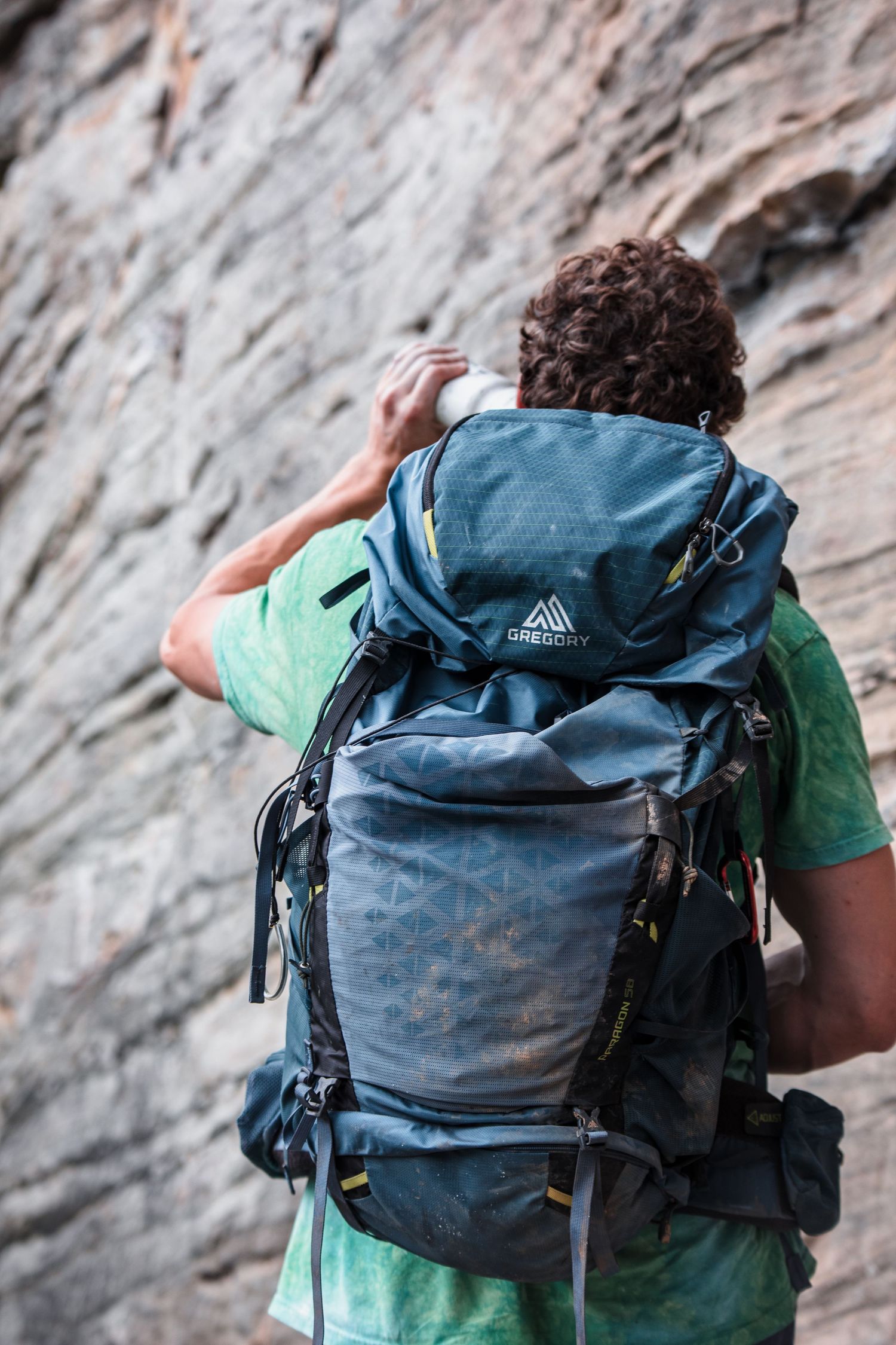 Rock climber wearing green shirt and blue backpack against steep cliff face.