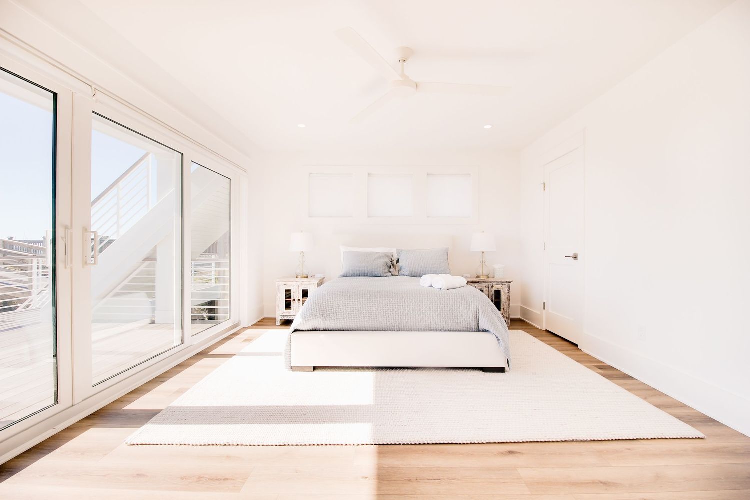 Bright minimalist bedroom with white walls, hardwood floors, gray bedding, and large sliding glass doors leading to a sunny balcony.