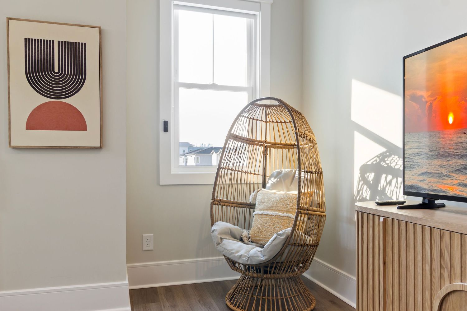 Modern boho bedroom corner with a wicker egg chair, abstract wall art, and a TV on a rattan stand.