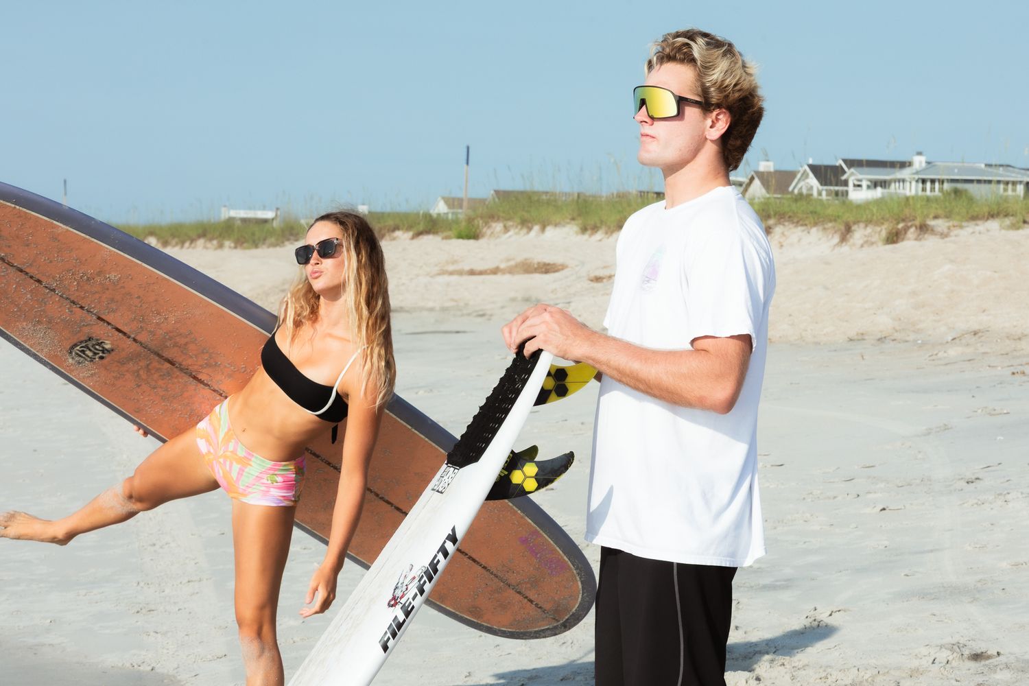Two young surfers on a sunny beach, girl in bikini and boy in white tee holding surfboards ready to surf.