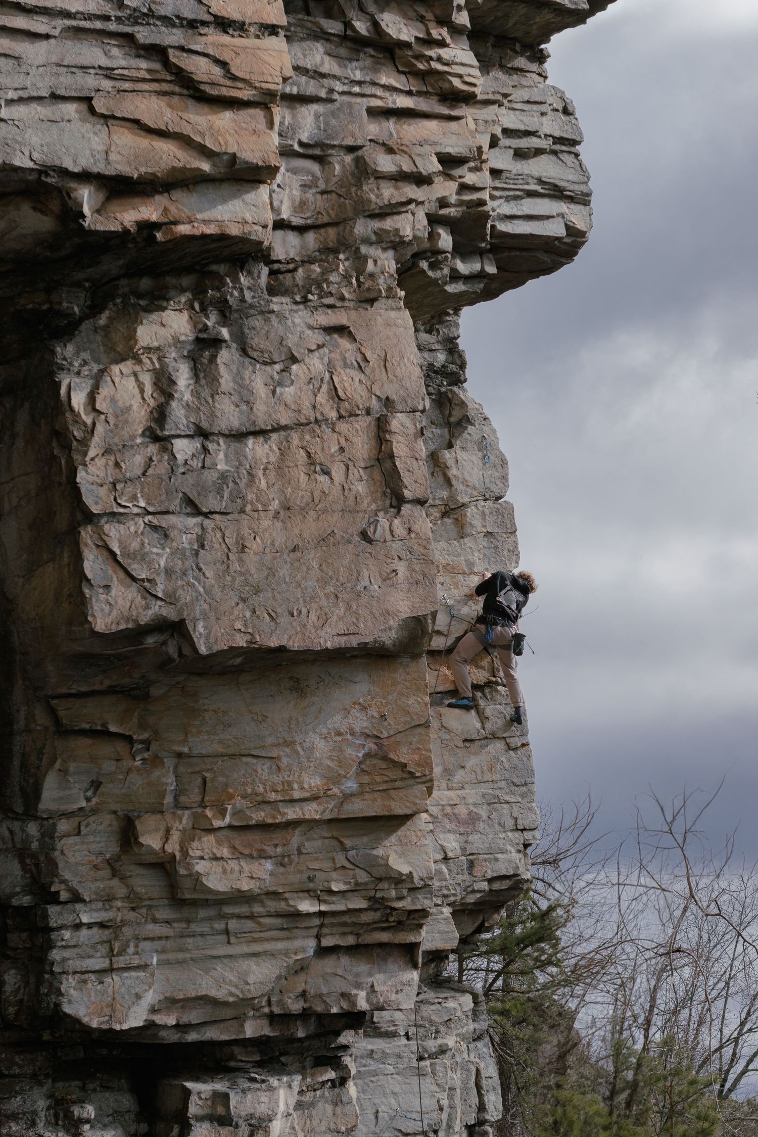 Rock climber scaling a massive layered sandstone cliff face under dramatic cloudy skies.
