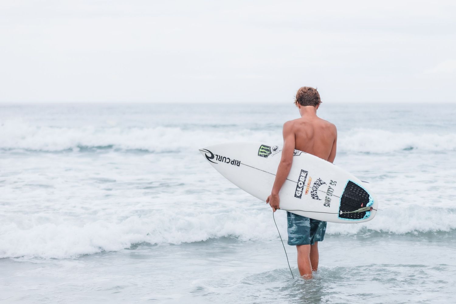 Surfer wading into ocean water carrying white surfboard on sunny day in Wrightsville Beach, NC.