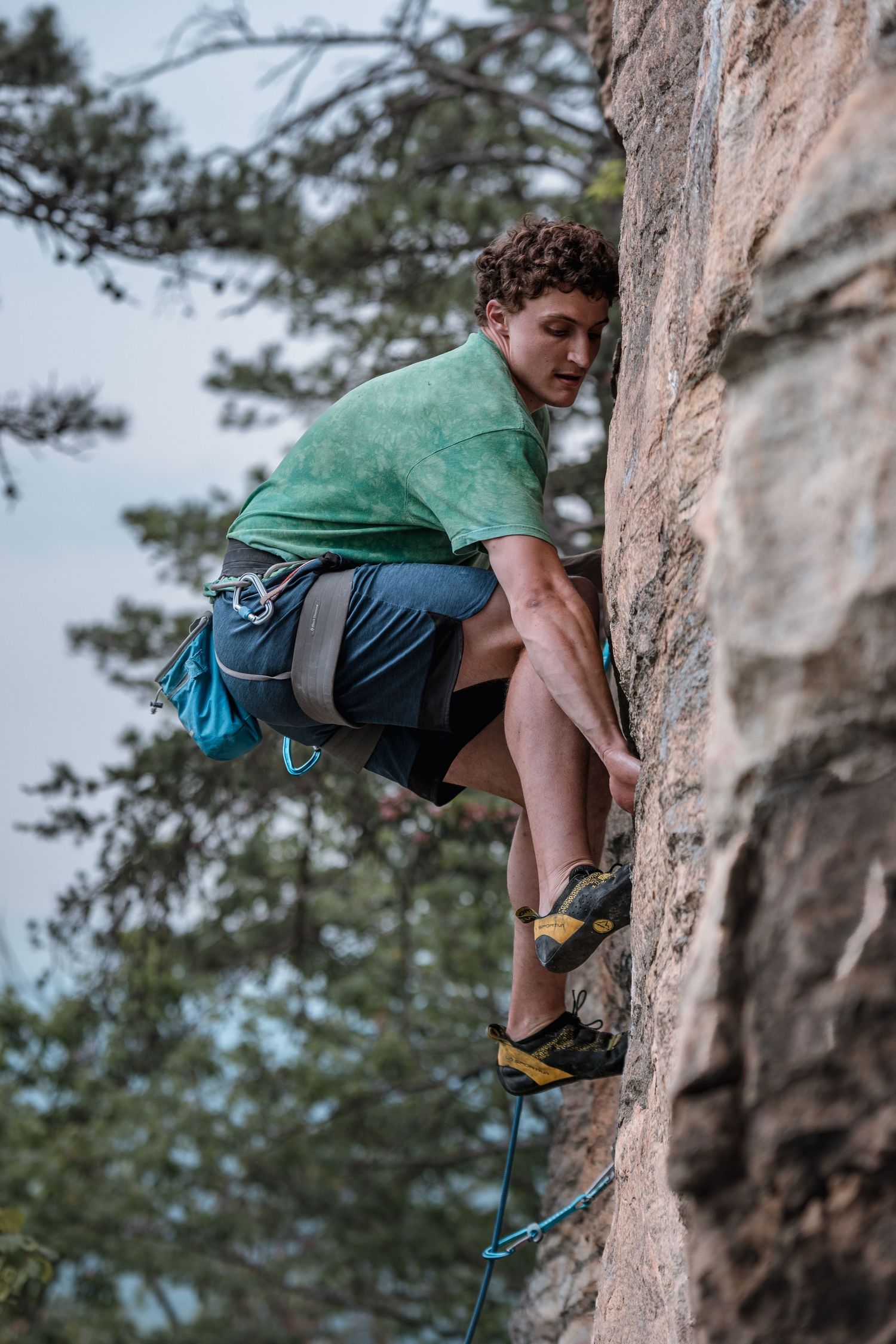 Young male rock climber scaling a steep cliff face wearing a green shirt and blue shorts outdoors.