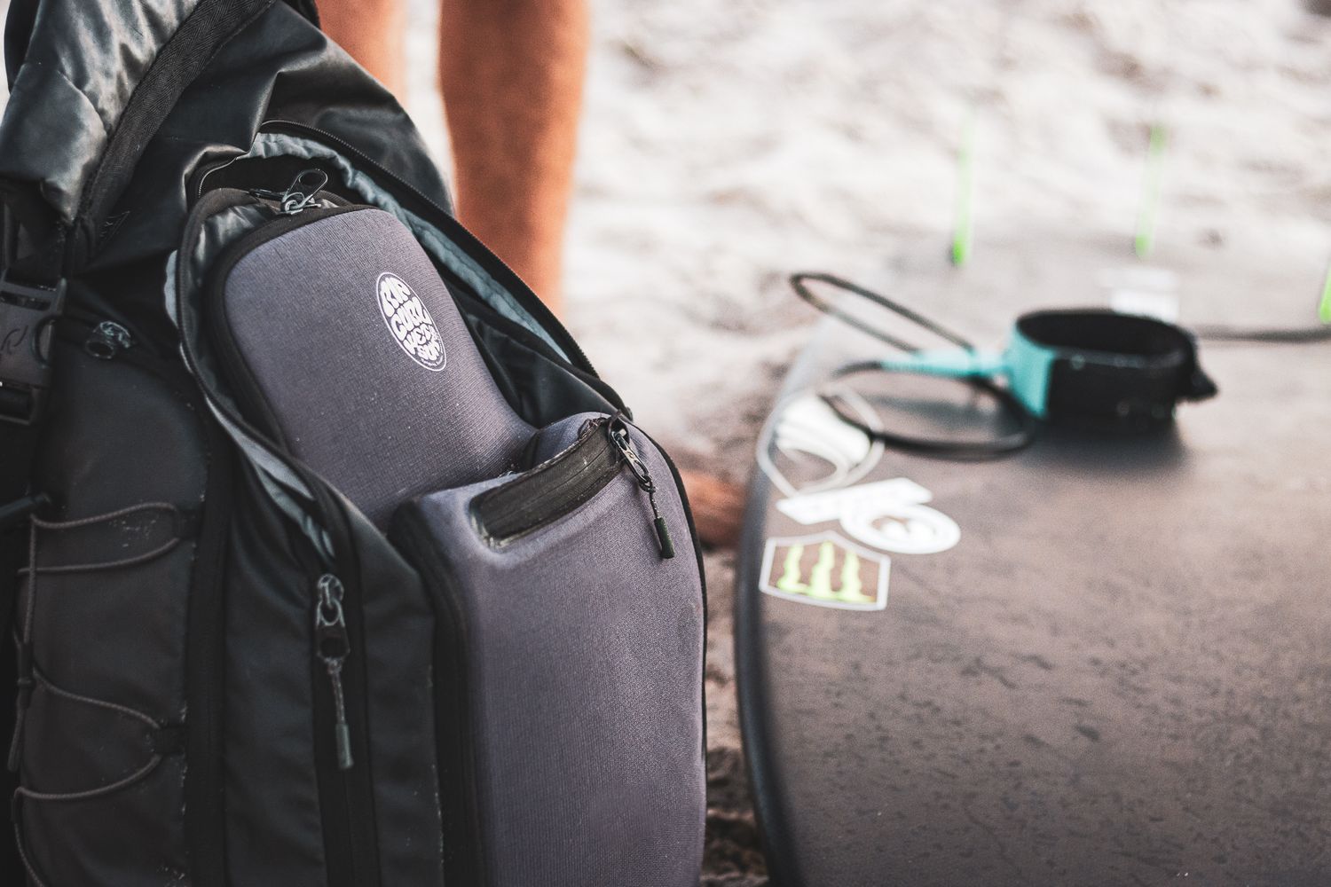 Close-up of a black backpack with side pocket, sunglasses, and accessories on sandy beach ground.