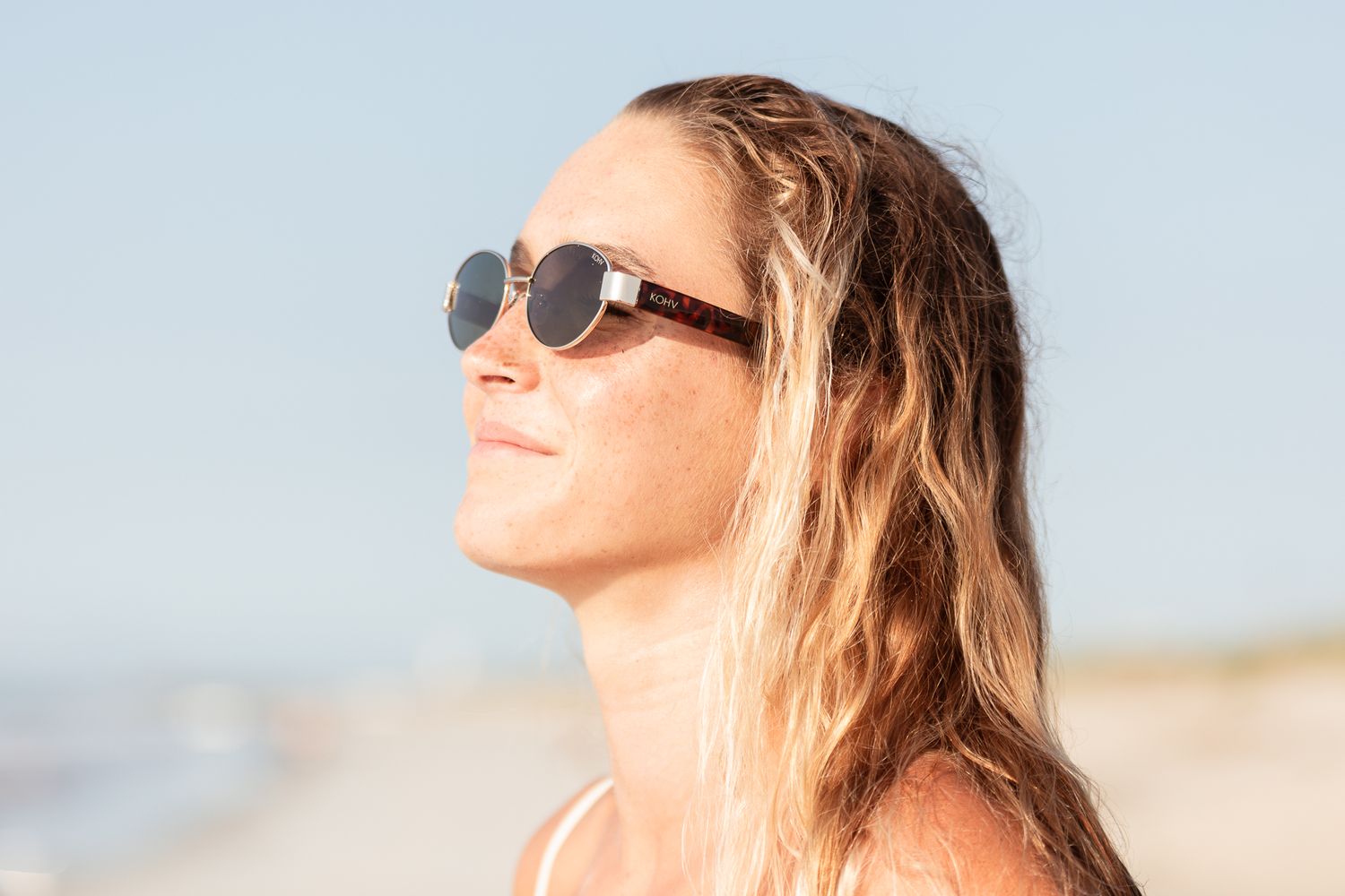 Person with wet blonde hair and sunglasses enjoying sunlight at the beach.