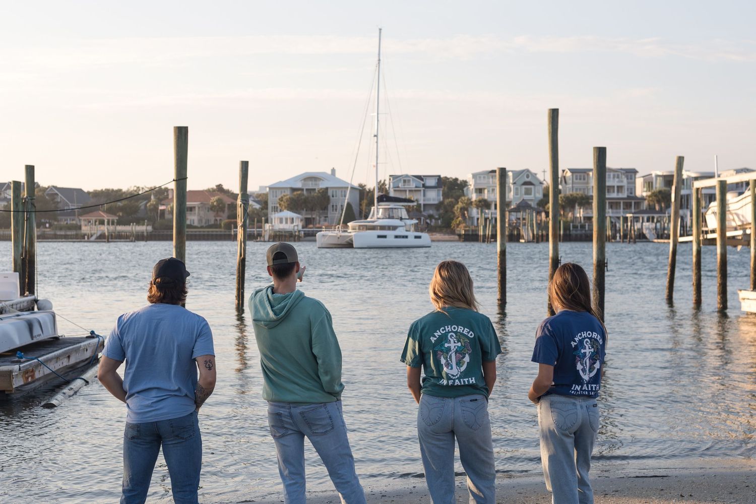 Four friends wearing matching sweatshirts stand on a dock overlooking boats and a marina at sunset.