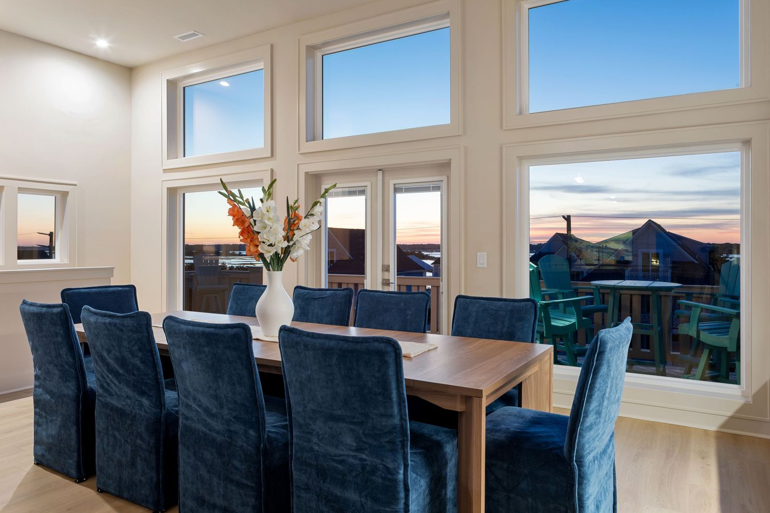 Elegant dining room with large windows offering sunset ocean views, featuring a wooden table and blue velvet chairs.