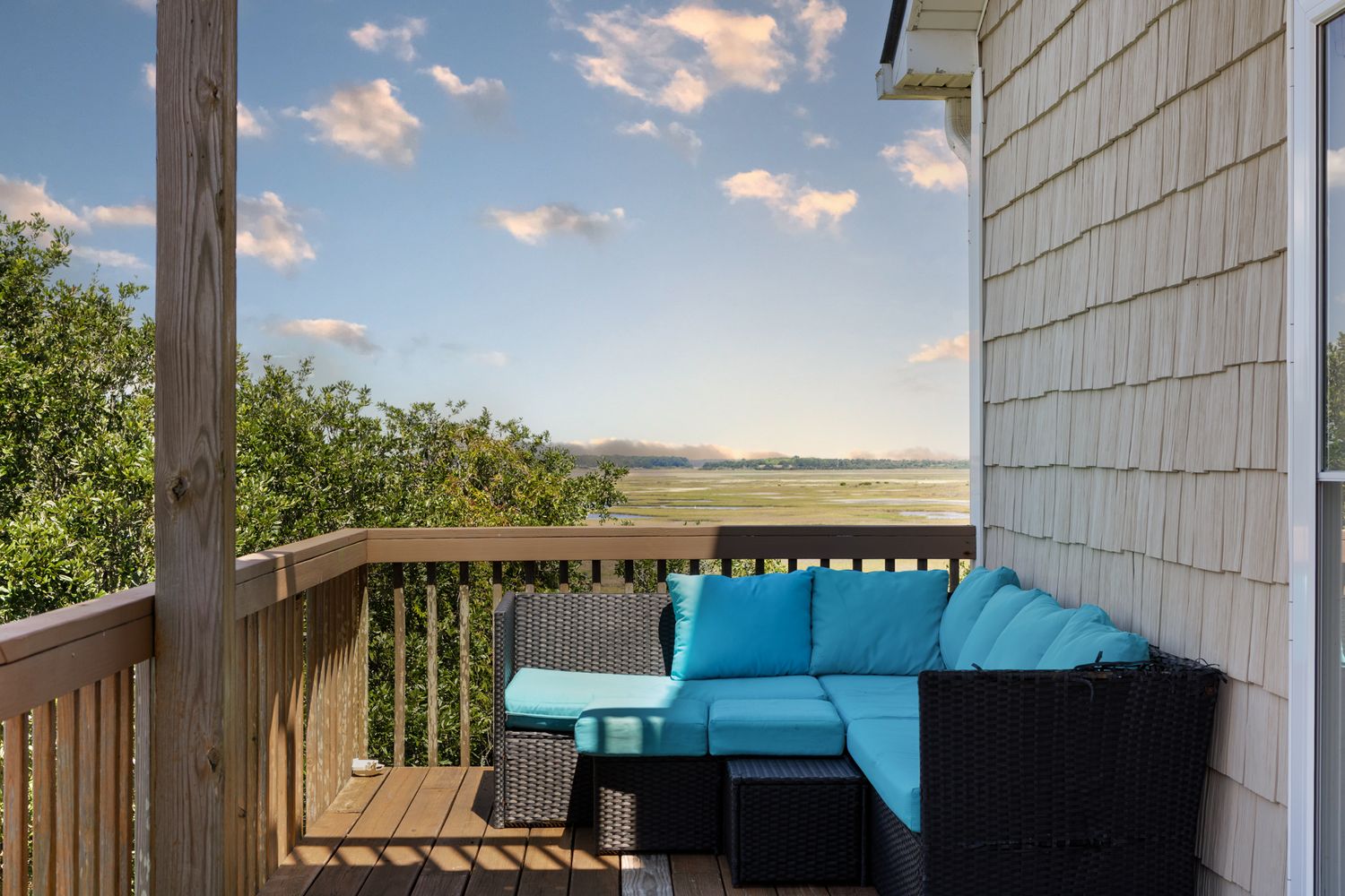 Outdoor deck with teal sectional sofa overlooking scenic marsh and blue sky with clouds.