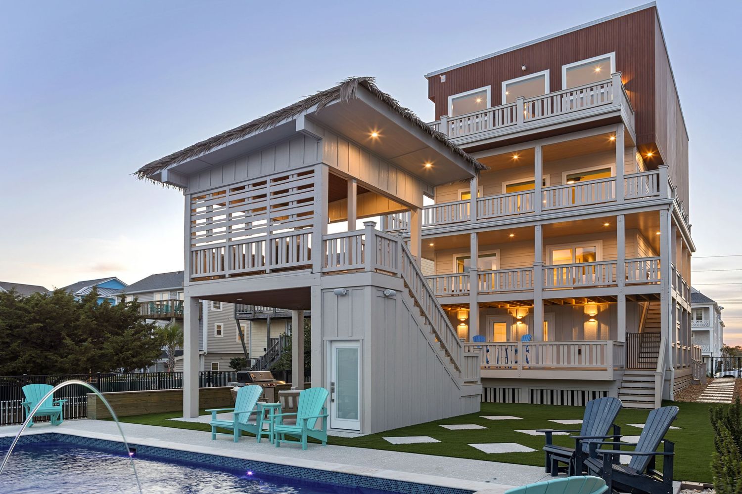 Multi-story beachside vacation home with wraparound decks, outdoor pool, and turquoise chairs at sunset.