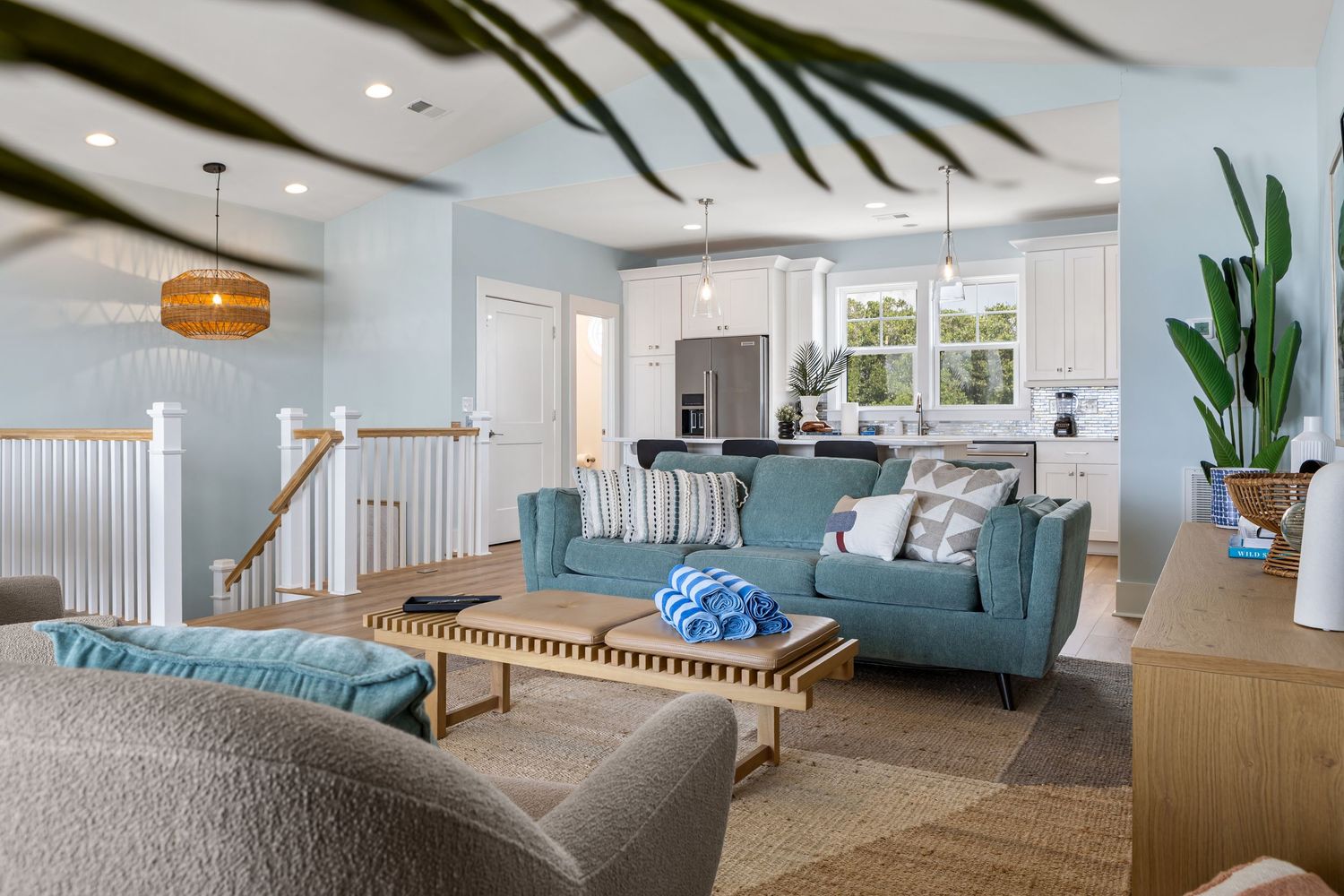 Bright coastal living room with blue sofas, woven coffee table, light blue walls, and open kitchen in background.