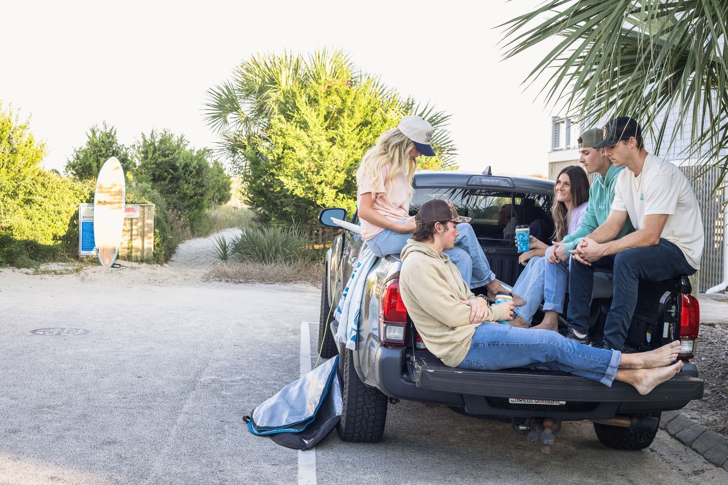 Four friends relaxing in the bed of a pickup truck near a beach parking lot on a sunny day.