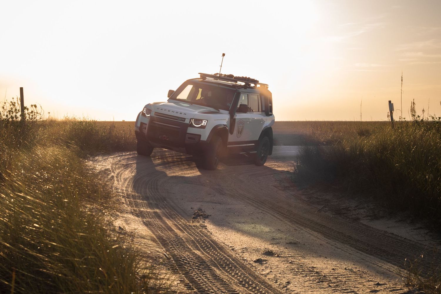 A white off road vehicle driving on a sand road at sunset creating a golden silhouette against the sky.