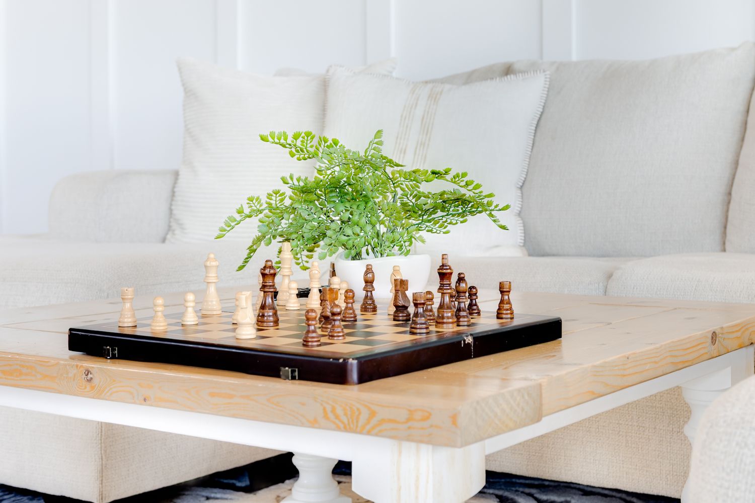 Chess board with wooden pieces and a green fern plant on a rustic wooden coffee table in a bright white living room.