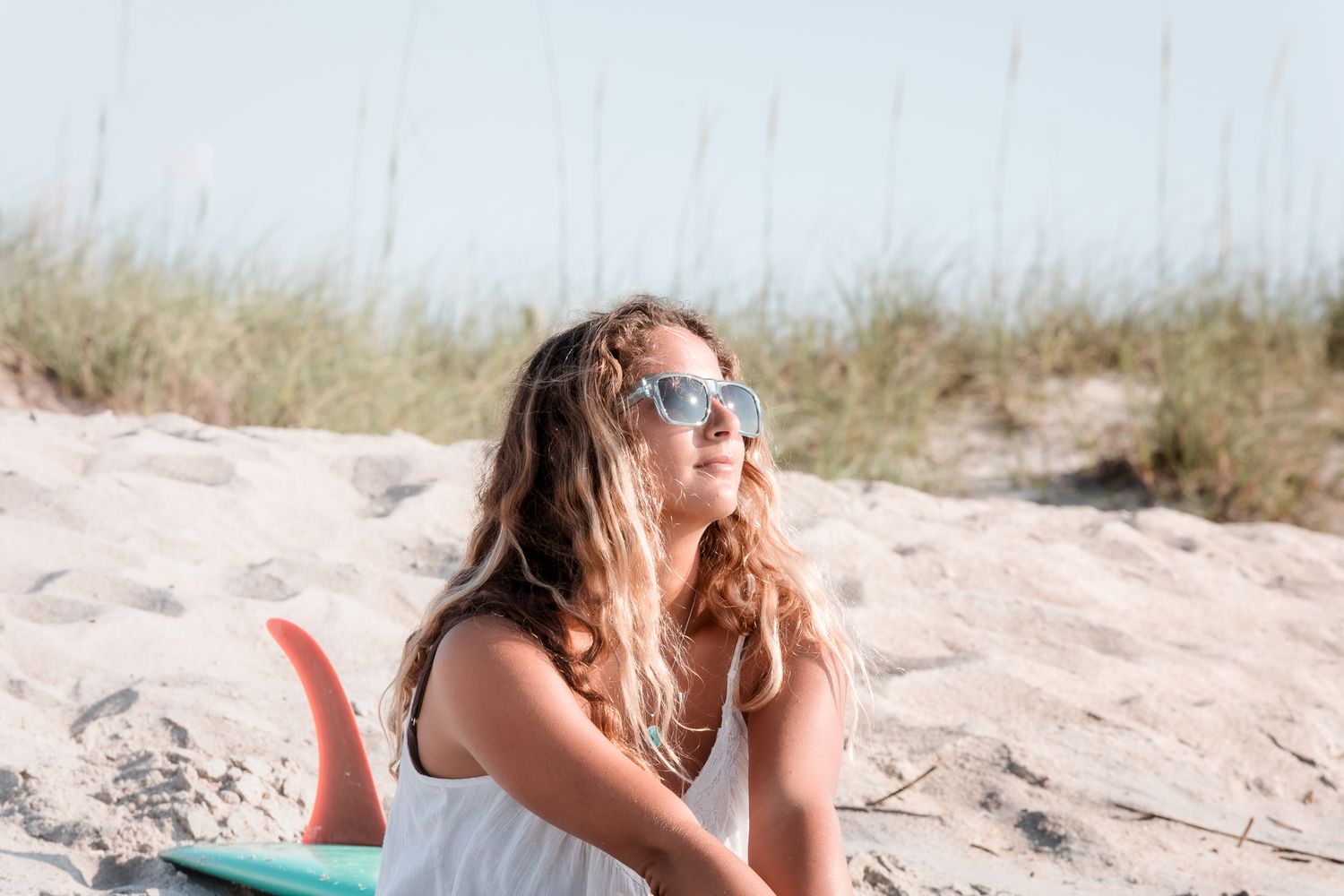 Person lying on beach sand wearing sunglasses and white dress on a sunny day.