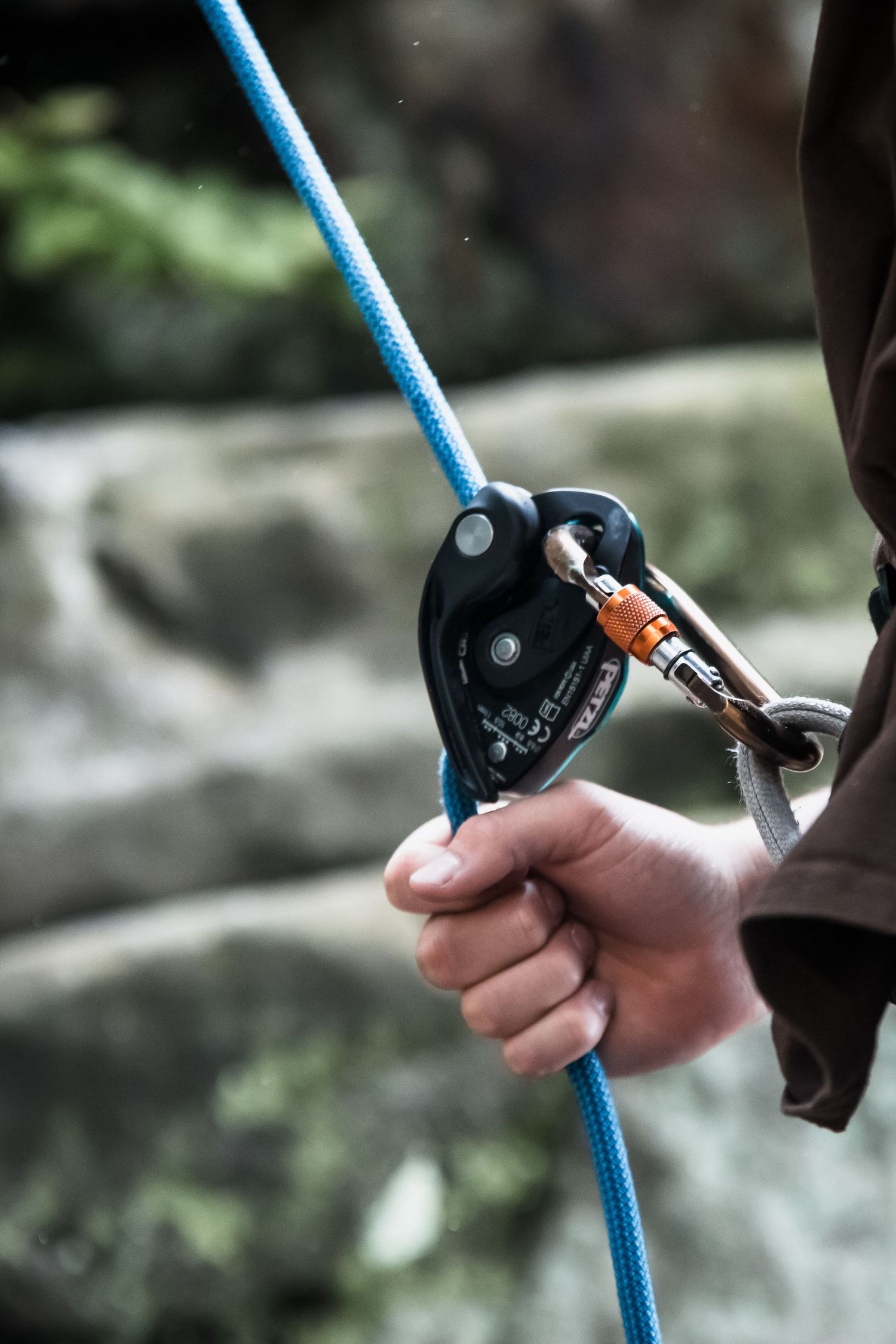 Close-up of a hand gripping a blue climbing rope with a black belay device and carabiner outdoors.