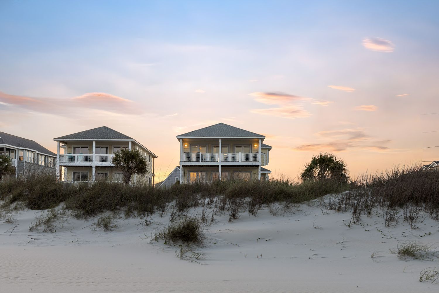 Beachfront vacation homes with wrap-around porches sit behind white sand dunes at sunset on a serene coastal shoreline.