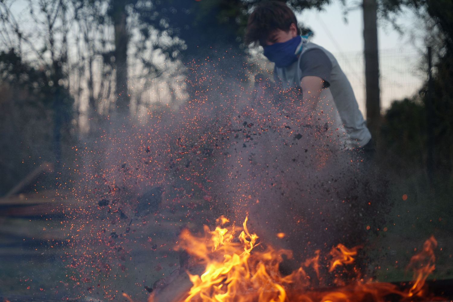 Shows a young adult putting a piece of wood on a fire with embers and ash being blown into the air