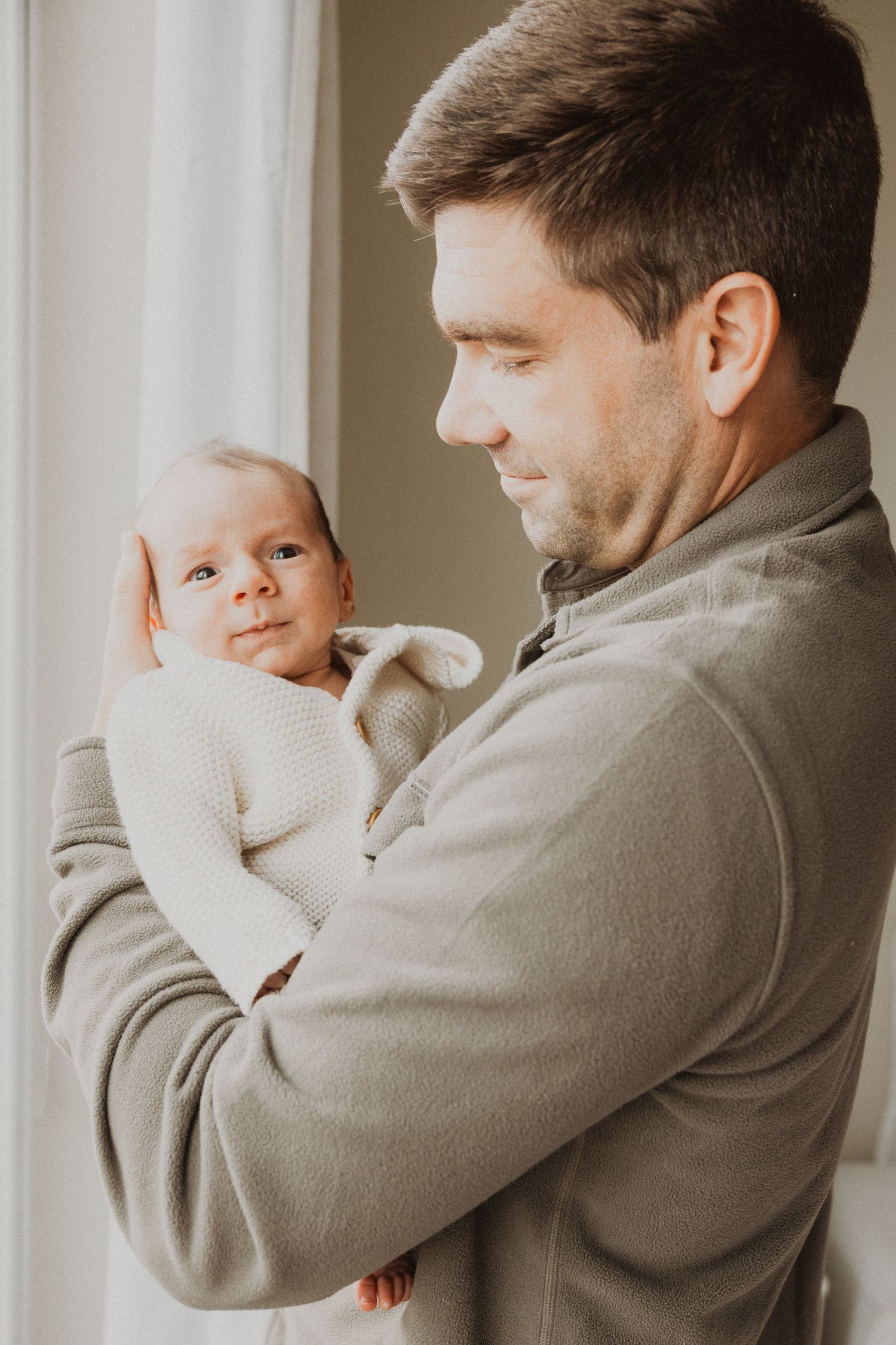 A father tenderly holds his newborn baby in a white knit outfit near a bright window, sharing a gentle moment.