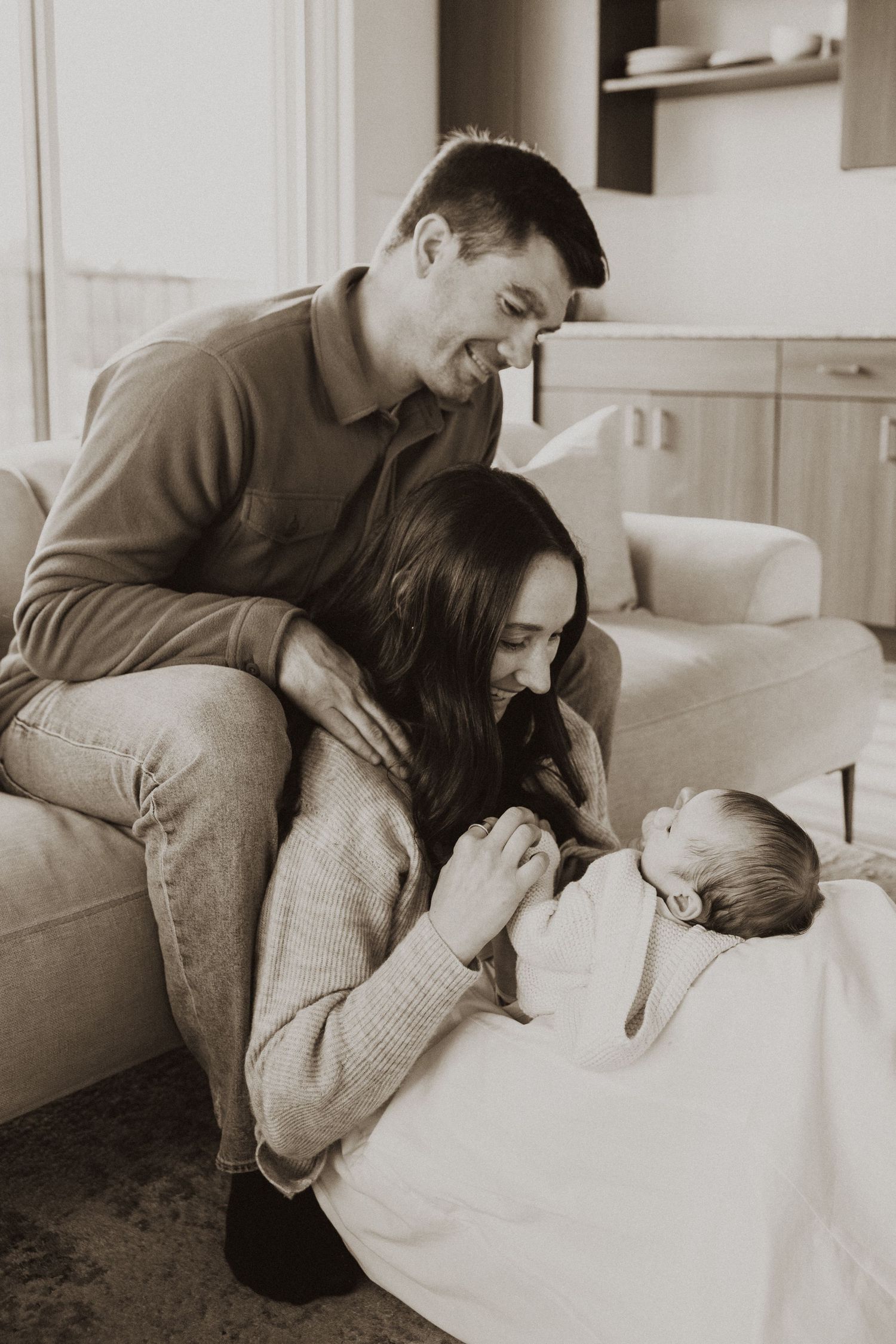 Black and white photo of new parents smiling at their newborn baby wrapped in a blanket at home.