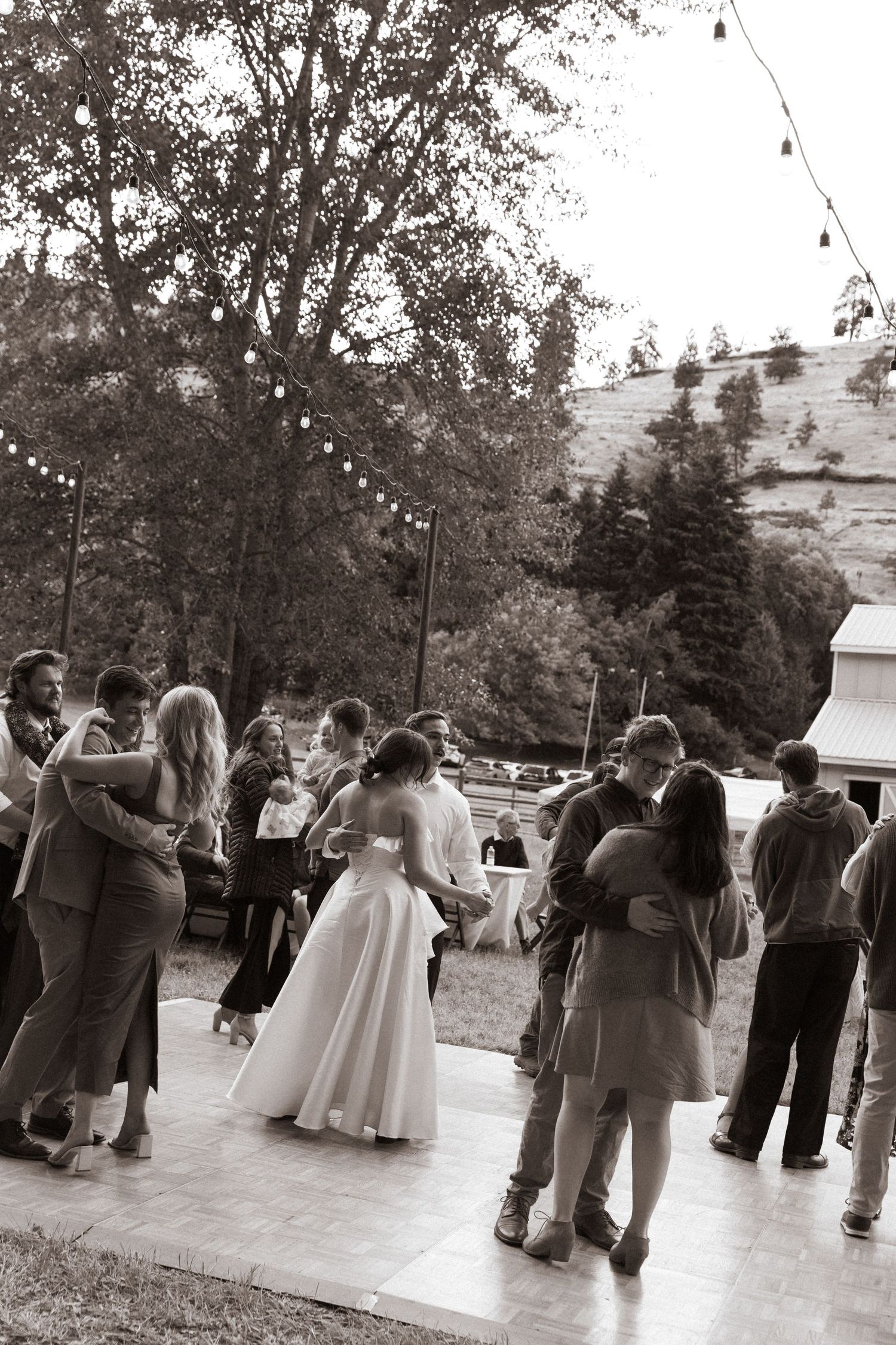 Black and white photo of wedding guests dancing outdoors under string lights with bride and groom at reception.