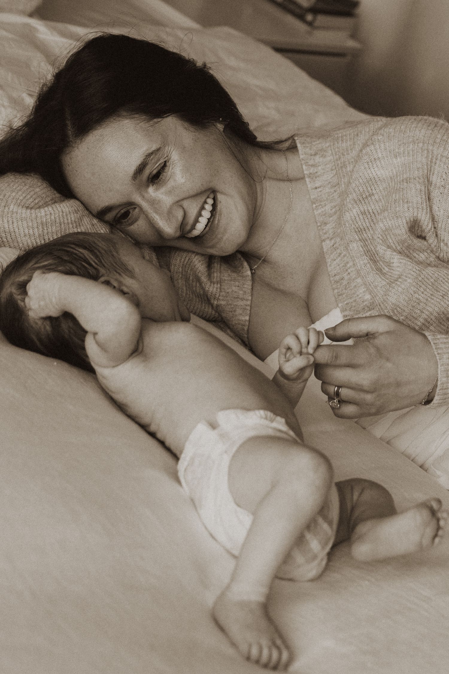 Smiling mother lying on bed with newborn baby in diaper, holding hands in sepia-toned photo.