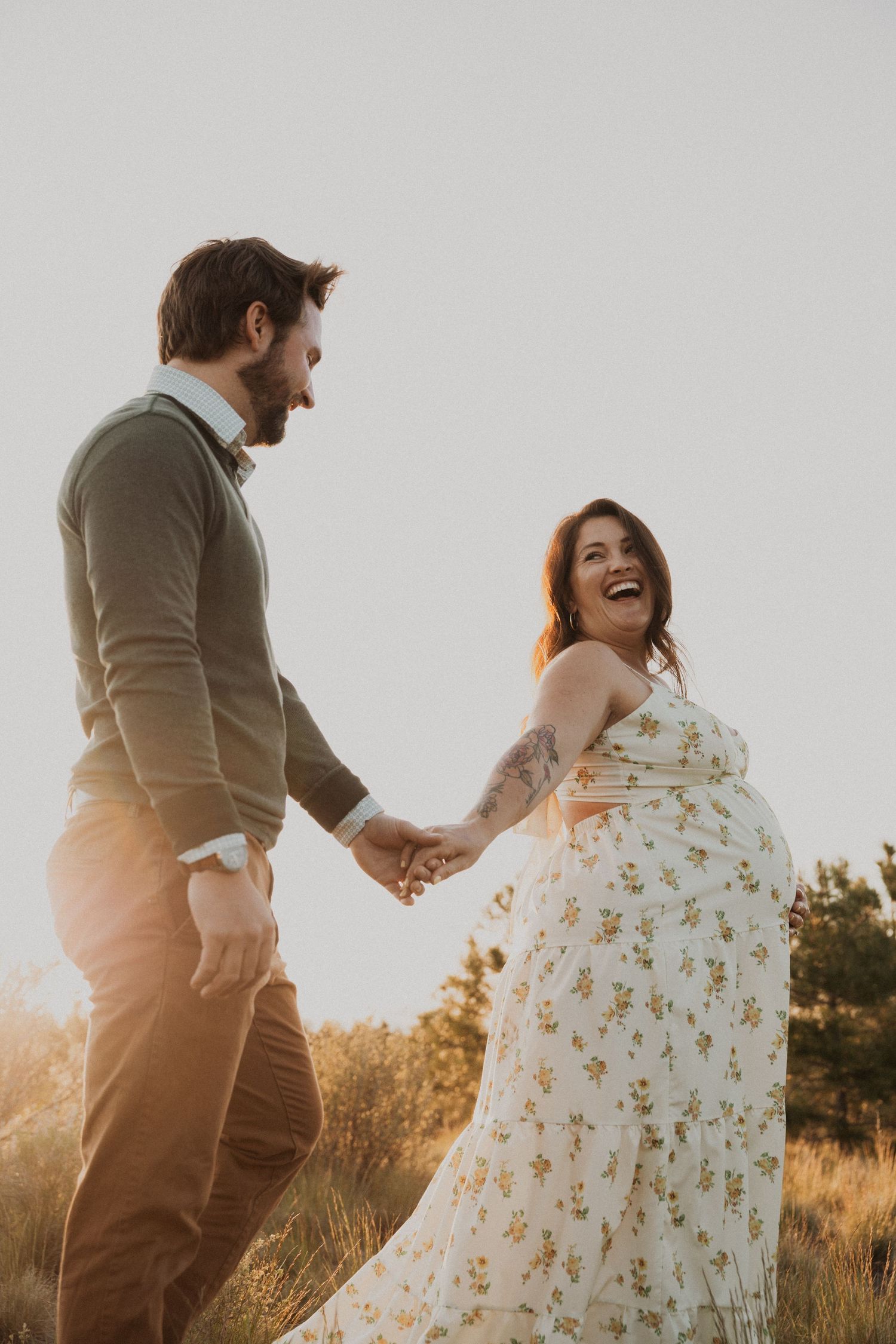 A laughing couple holds hands in a sunlit field, the woman in a floral dress with a baby bump, the man in a green sweater.