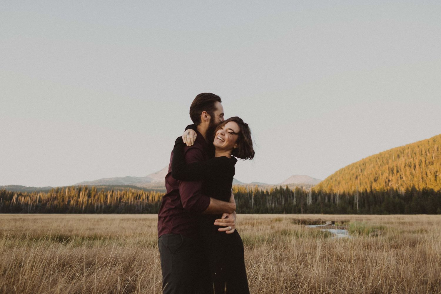 A romantic couple shares tender moments in a vast field with mountains on the horizon during sunset.