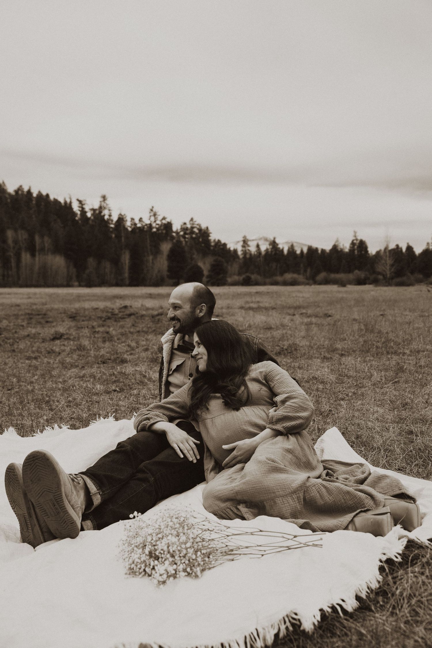 Expecting couple relaxes on a white blanket in an open field with wildflowers in a sepia-toned photo.