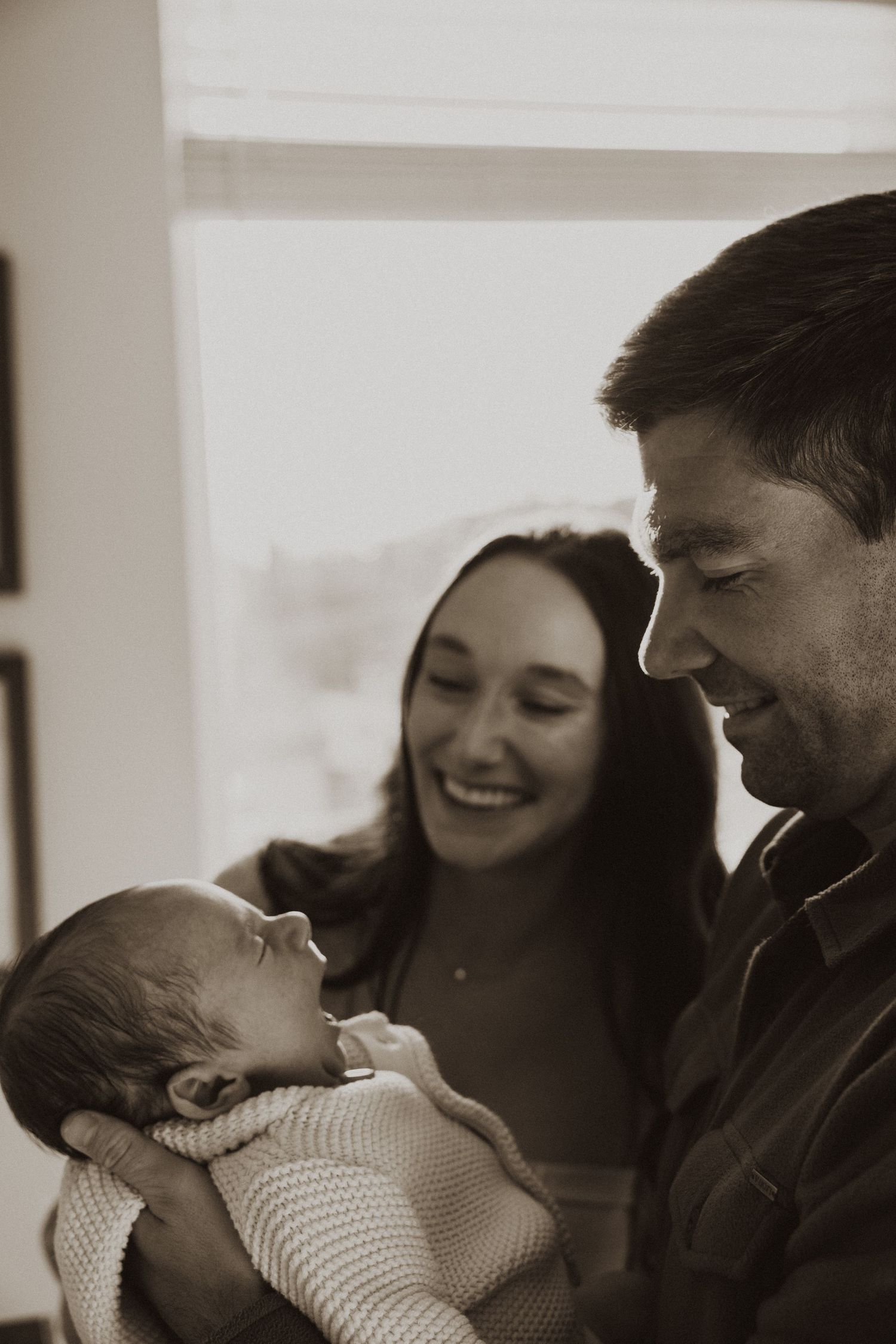 Black and white photo of smiling parents lovingly gazing at their newborn baby wrapped in a white blanket.