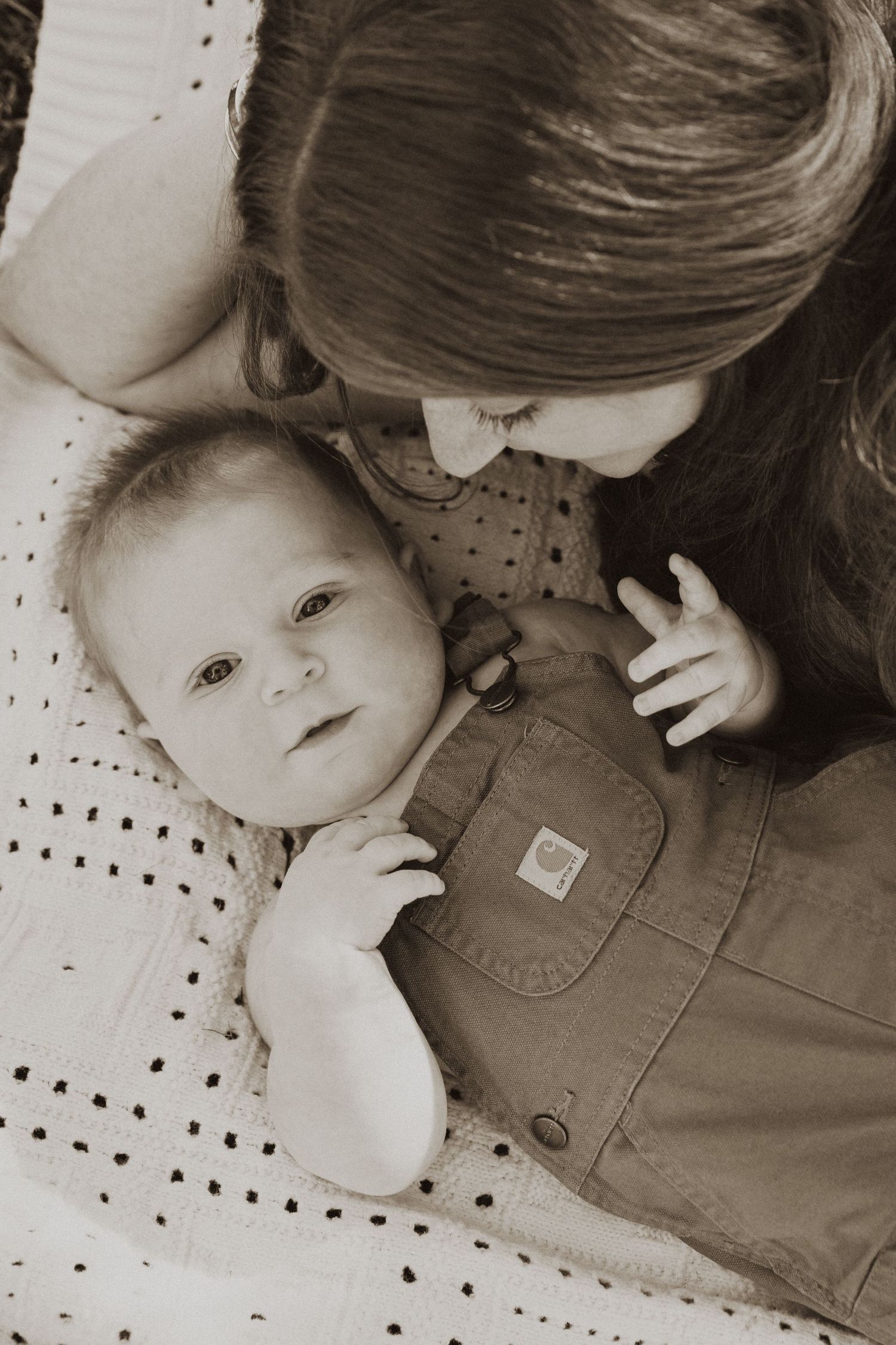 Sepia-toned photo of a mother lovingly gazing at her baby in overalls lying on a quilted blanket.