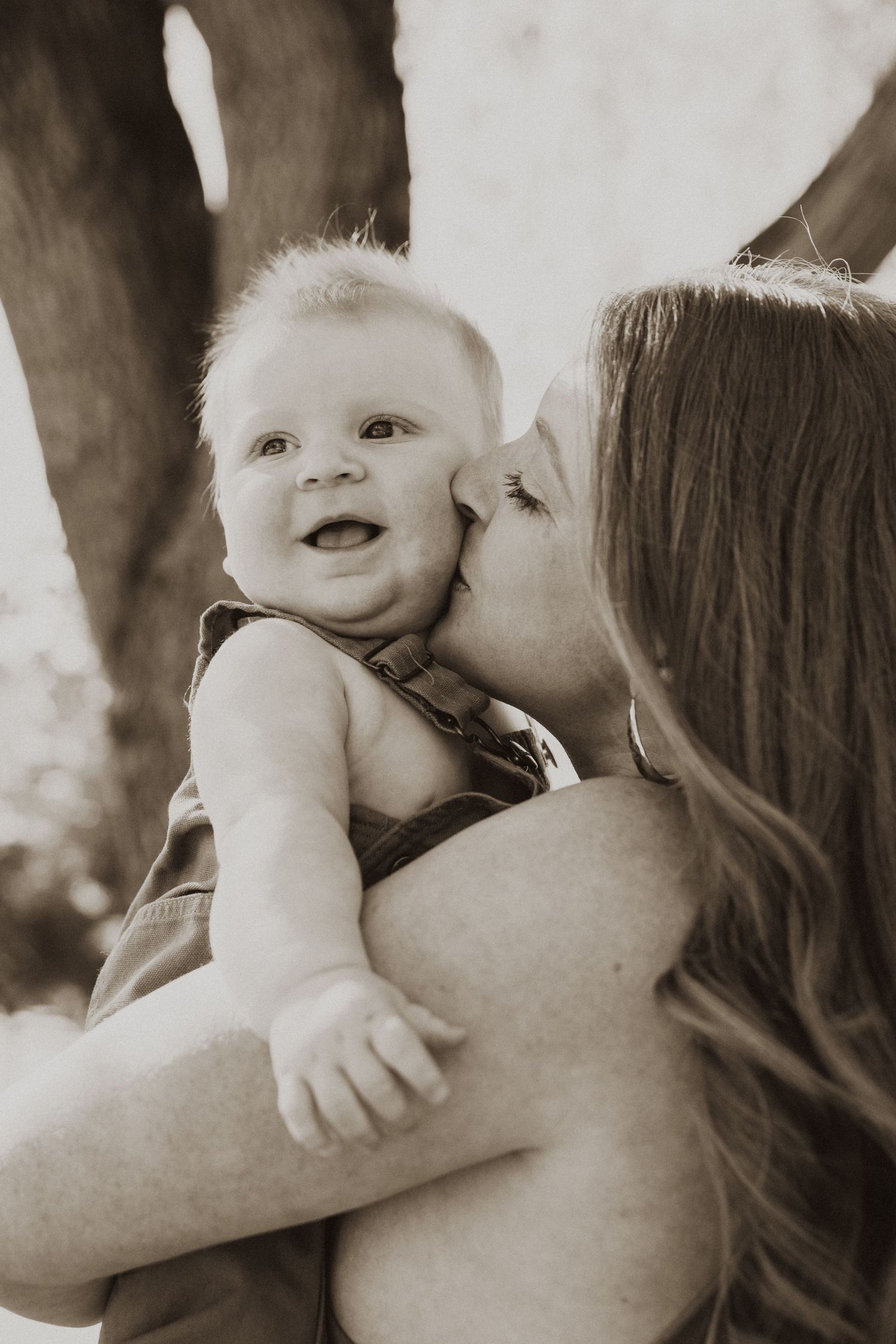A mother tenderly kisses her smiling baby outdoors in a warm sepia-toned portrait.