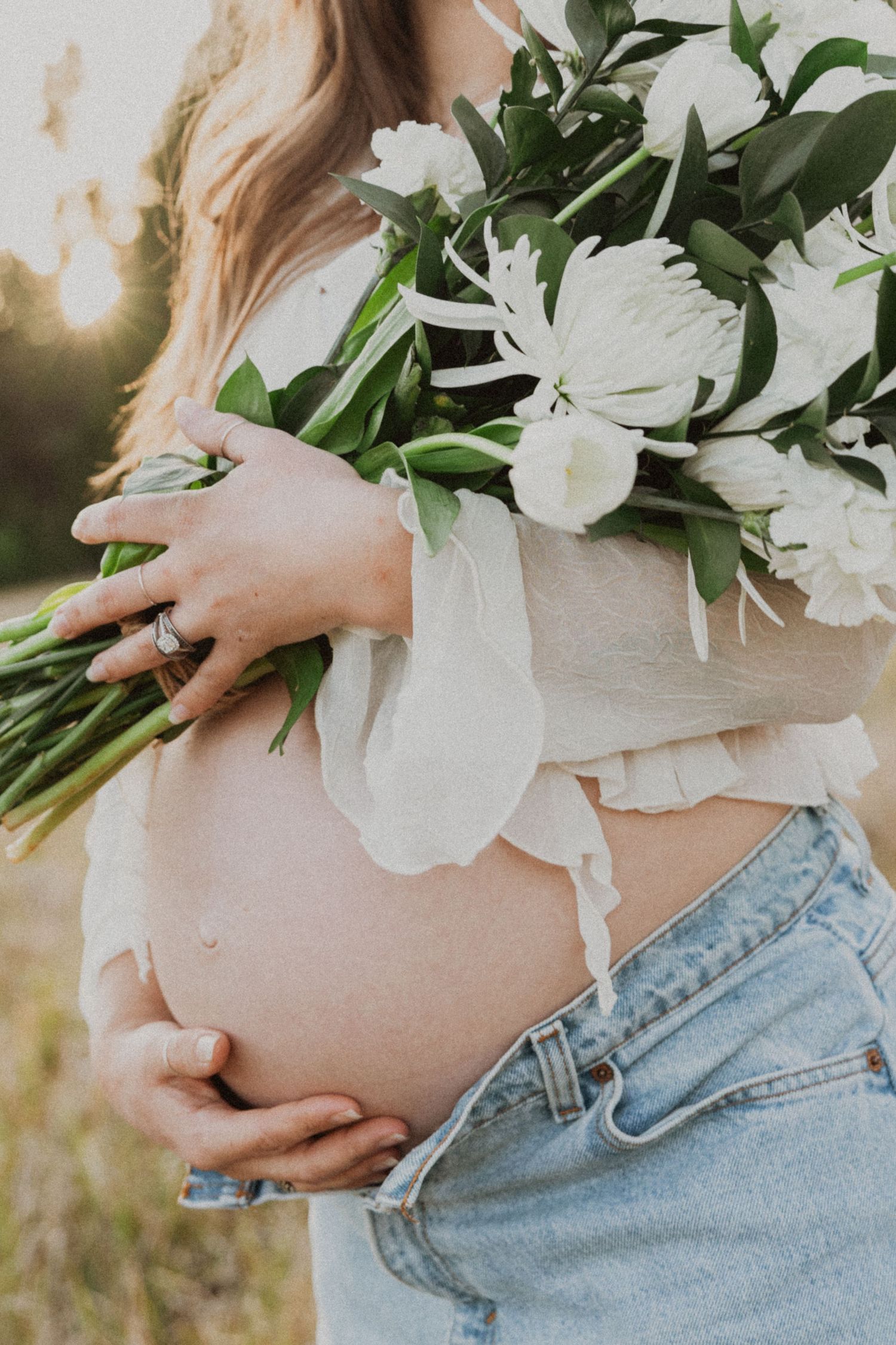 Pregnant woman in white top and jeans holding bouquet of white flowers over baby bump.