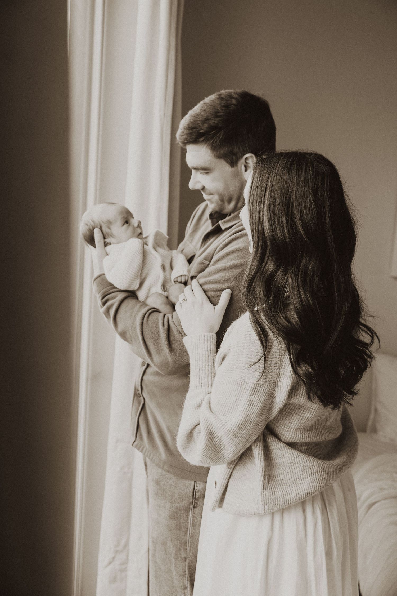 Sepia-toned photo of new parents standing by a window, father holding newborn baby while mother looks on lovingly.
