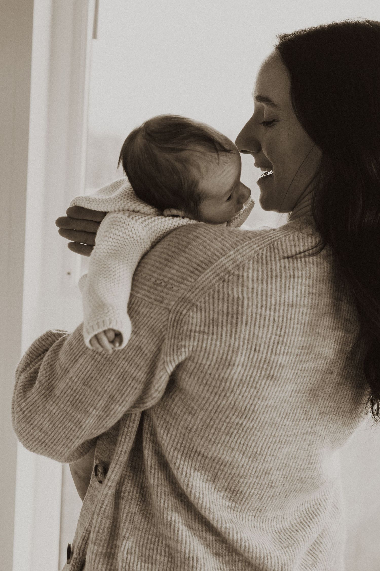 A mother smiling warmly while holding her newborn baby in a soft sepia-toned portrait photo.