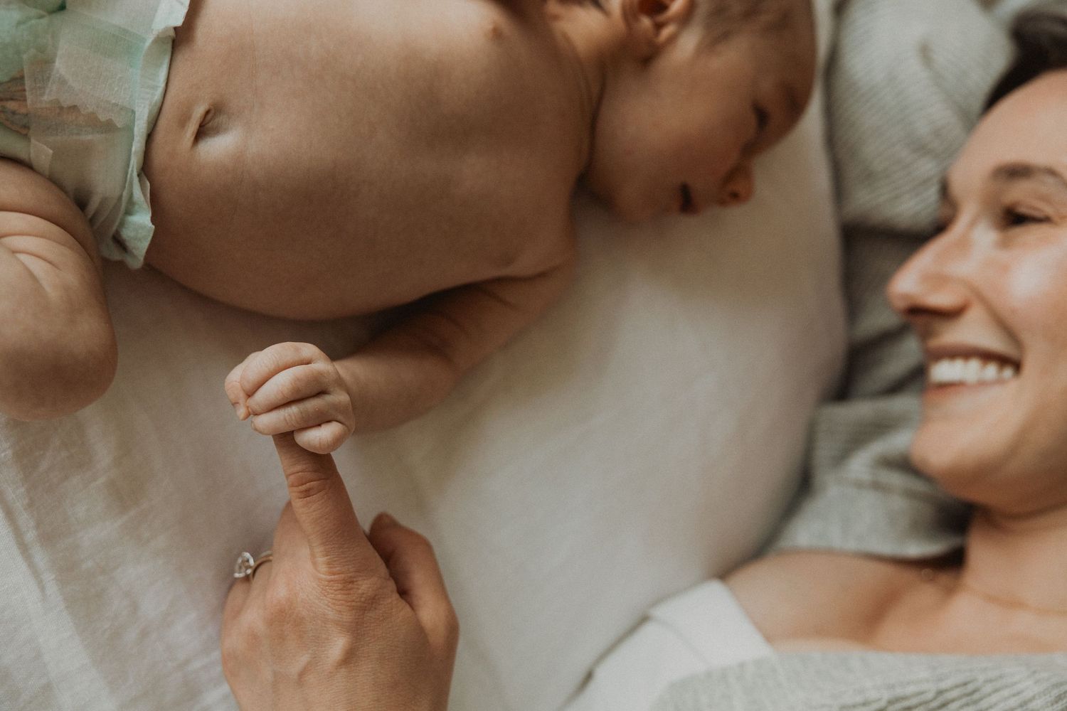 Mother smiling while lying next to sleeping newborn baby holding her finger on white bedding.