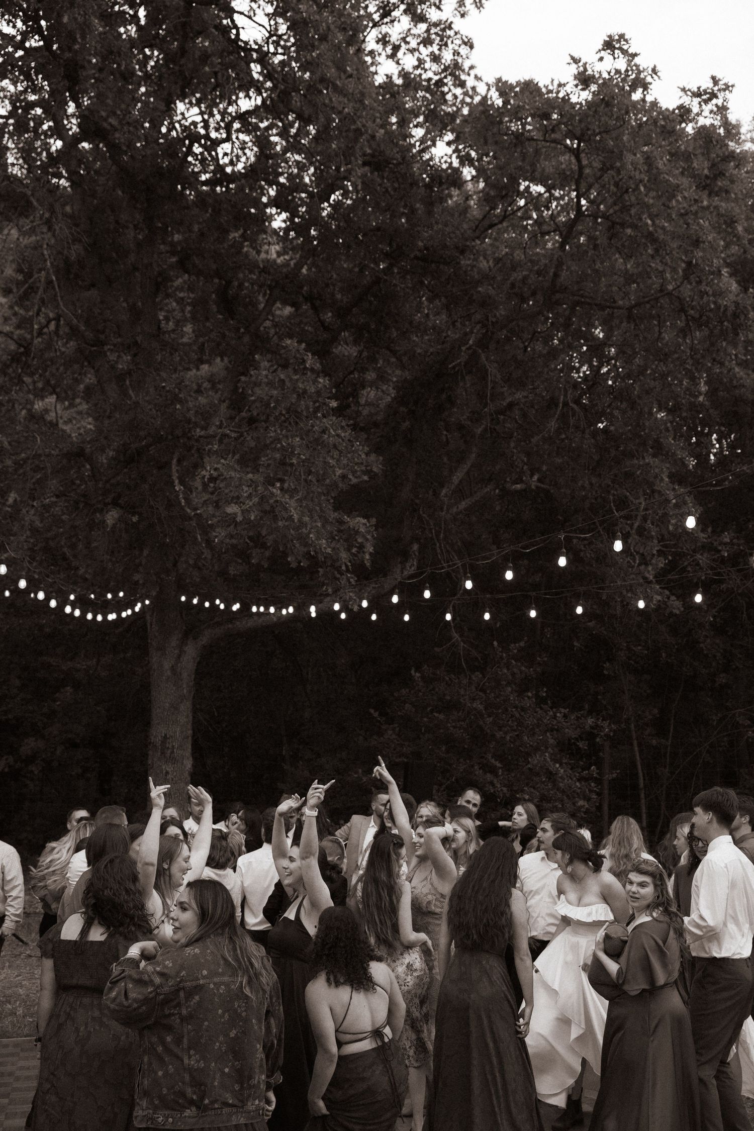 Wedding guests dance outdoors under string lights at night surrounded by trees in a black and white reception photo.