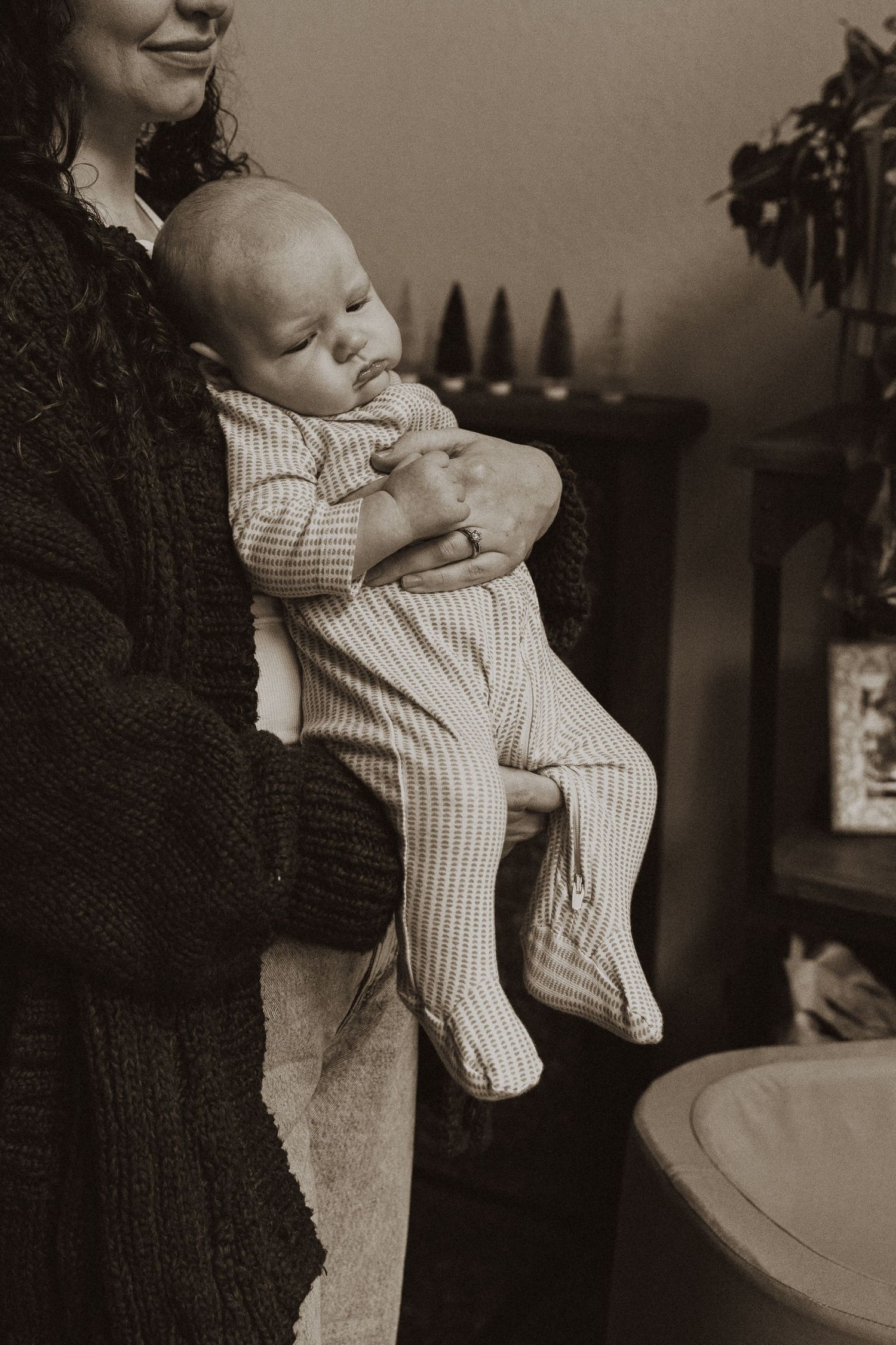 Mother in brown knit cardigan cradling sleeping newborn baby in sepia-toned indoor portrait.