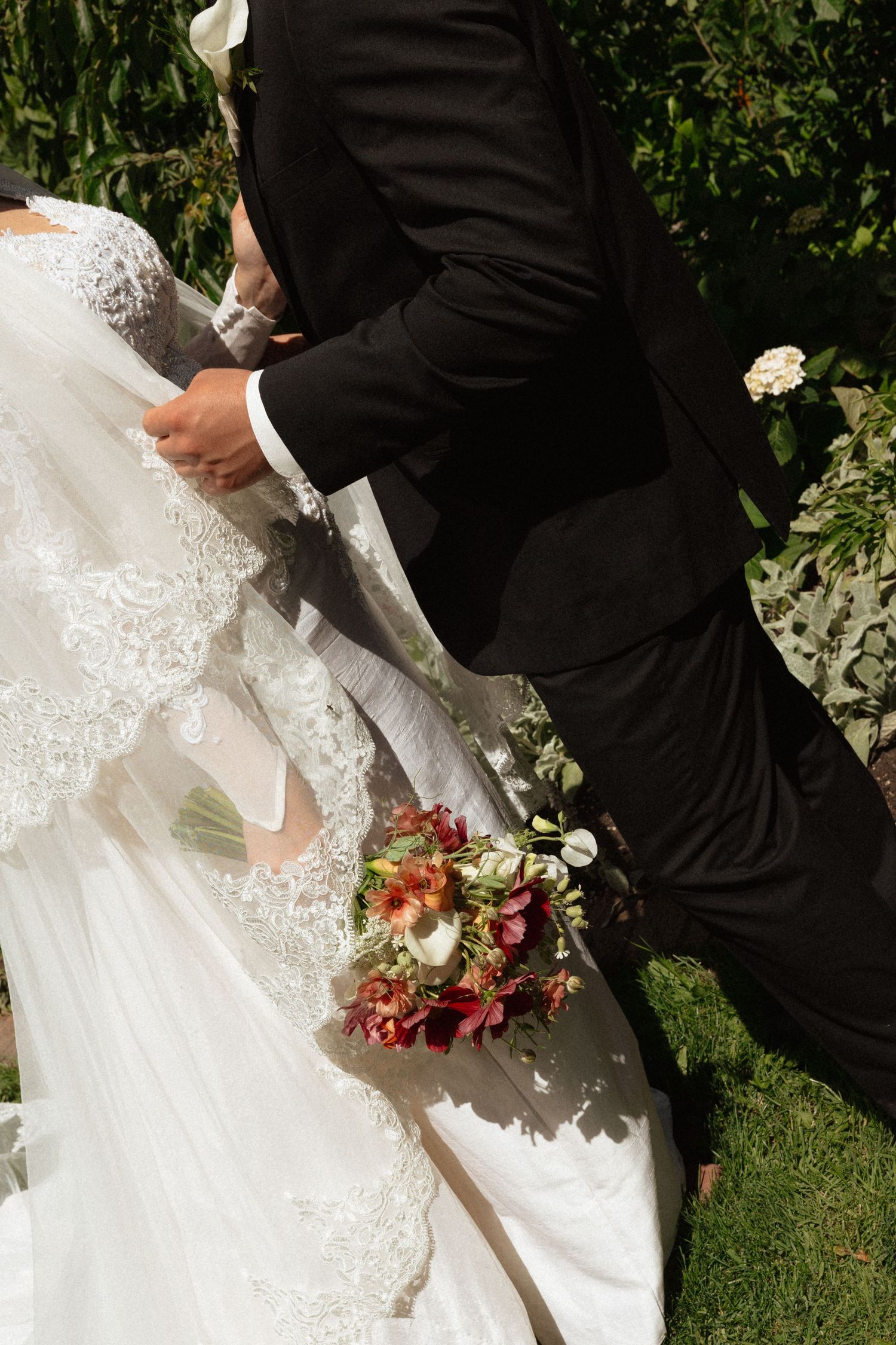 Bride and groom walking hand in hand, bride holding a red and white floral bouquet in a lace gown.