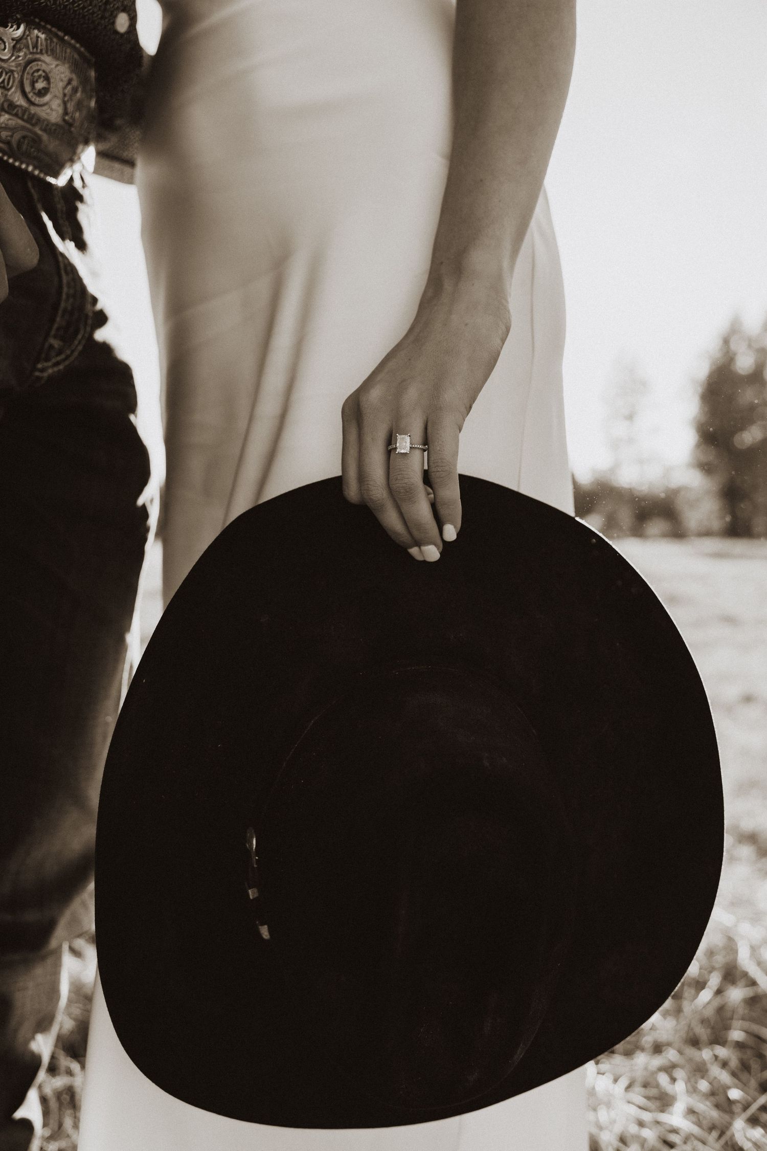 Sepia-toned photo of a bride holding a black cowboy hat, wearing an engagement ring, standing beside a groom in jeans.