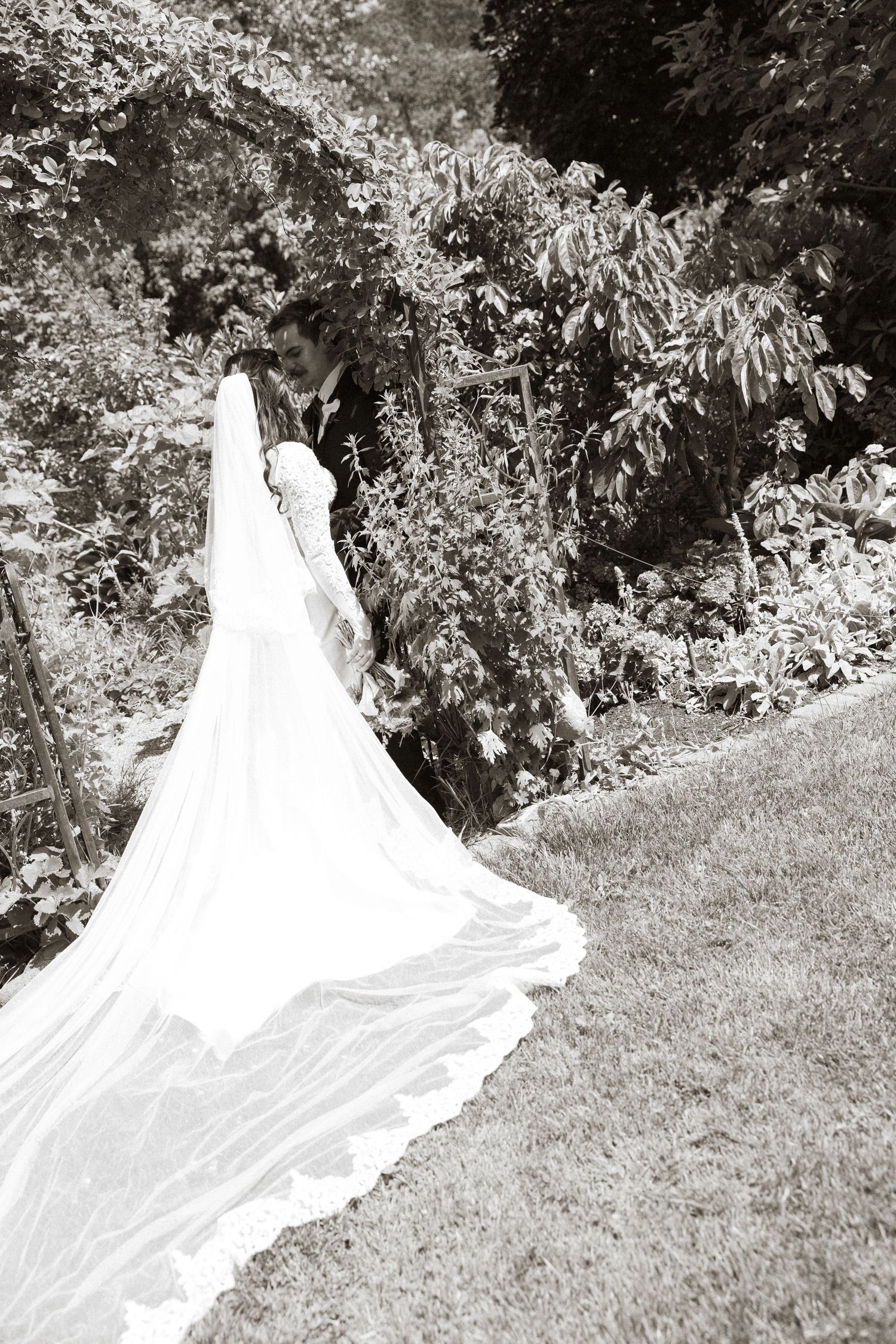 Bride in a stunning white gown with a long flowing train walks with groom through a lush garden in black and white.